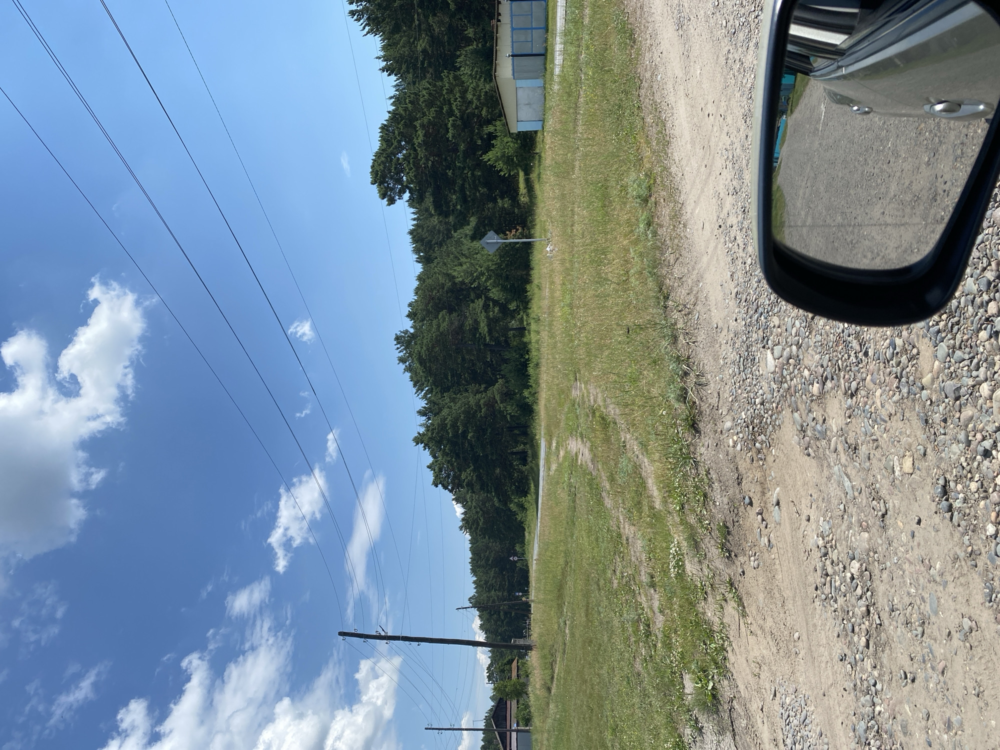 The image depicts a rural landscape under a bright, cloudy sky. In the foreground, a gravel and dirt road is visible, with tire tracks indicating recent vehicle passage. To the left, a grassy field with scattered wildflowers stretches towards a cluster of wooden buildings and power lines. A dirt path, also with tire tracks, branches off into the field. In the middle ground, a paved road curves through a field and disappears into a dense forest of pine trees. A single, diamond-shaped blue and white sign is posted in the field near the road. To the right, a light-colored building with a blue trim and a blue metal fence stands at the edge of the forest. The reflection in the car's side mirror shows the continuation of the gravel road. This scene is consistent with the outskirts of the village of Znamenka, Russia, characterized by its semi-rural setting with residential structures, unpaved roads, and surrounding natural vegetation.
