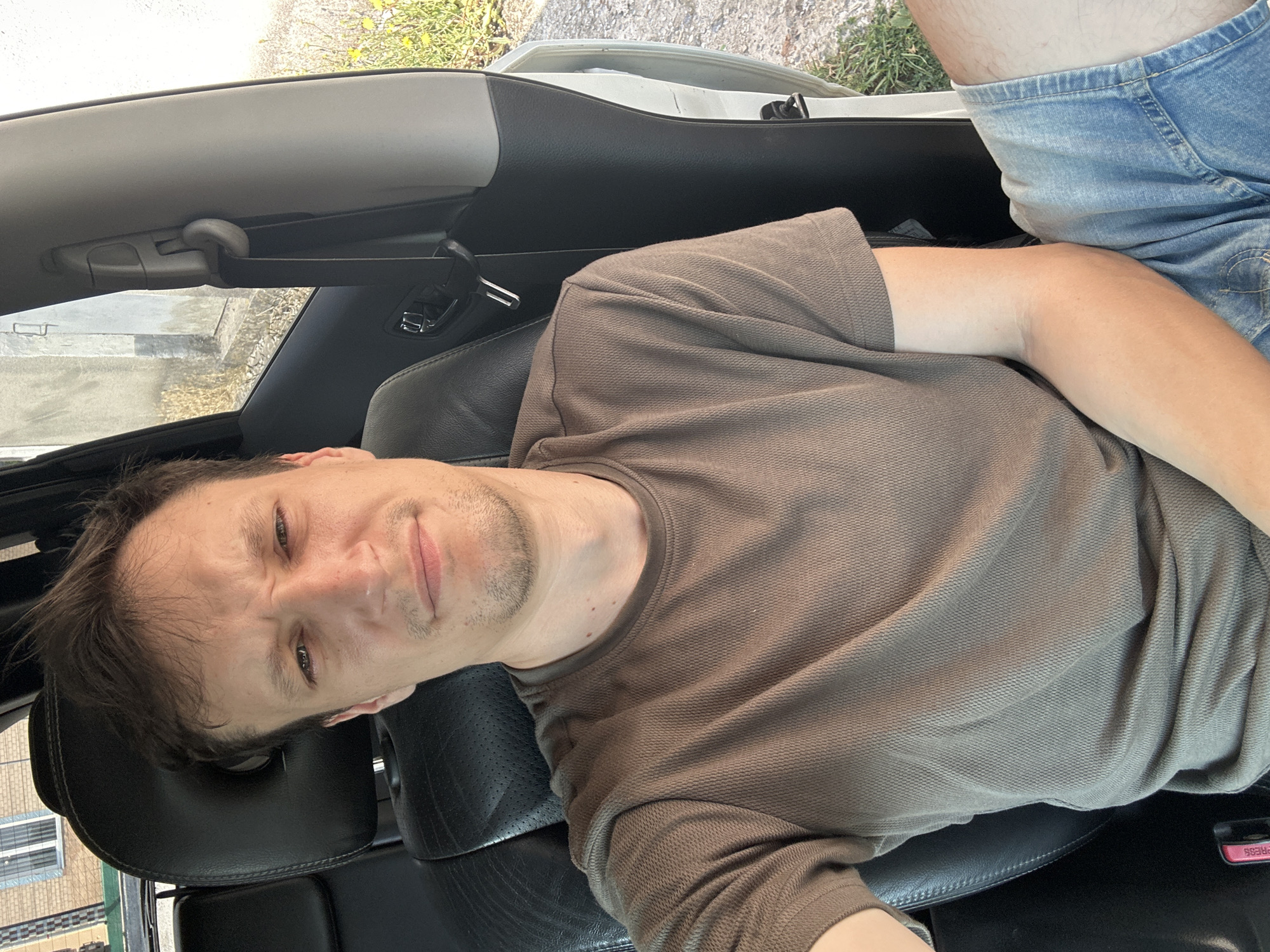 A man is sitting in the driver's seat of a car, taking a selfie. He has short brown hair and a stubble beard. He is wearing a brown t-shirt and blue denim shorts. The car appears to be parked on a gravel road with some vegetation and buildings visible in the background. The location is the city of Shatava, Ukraine.