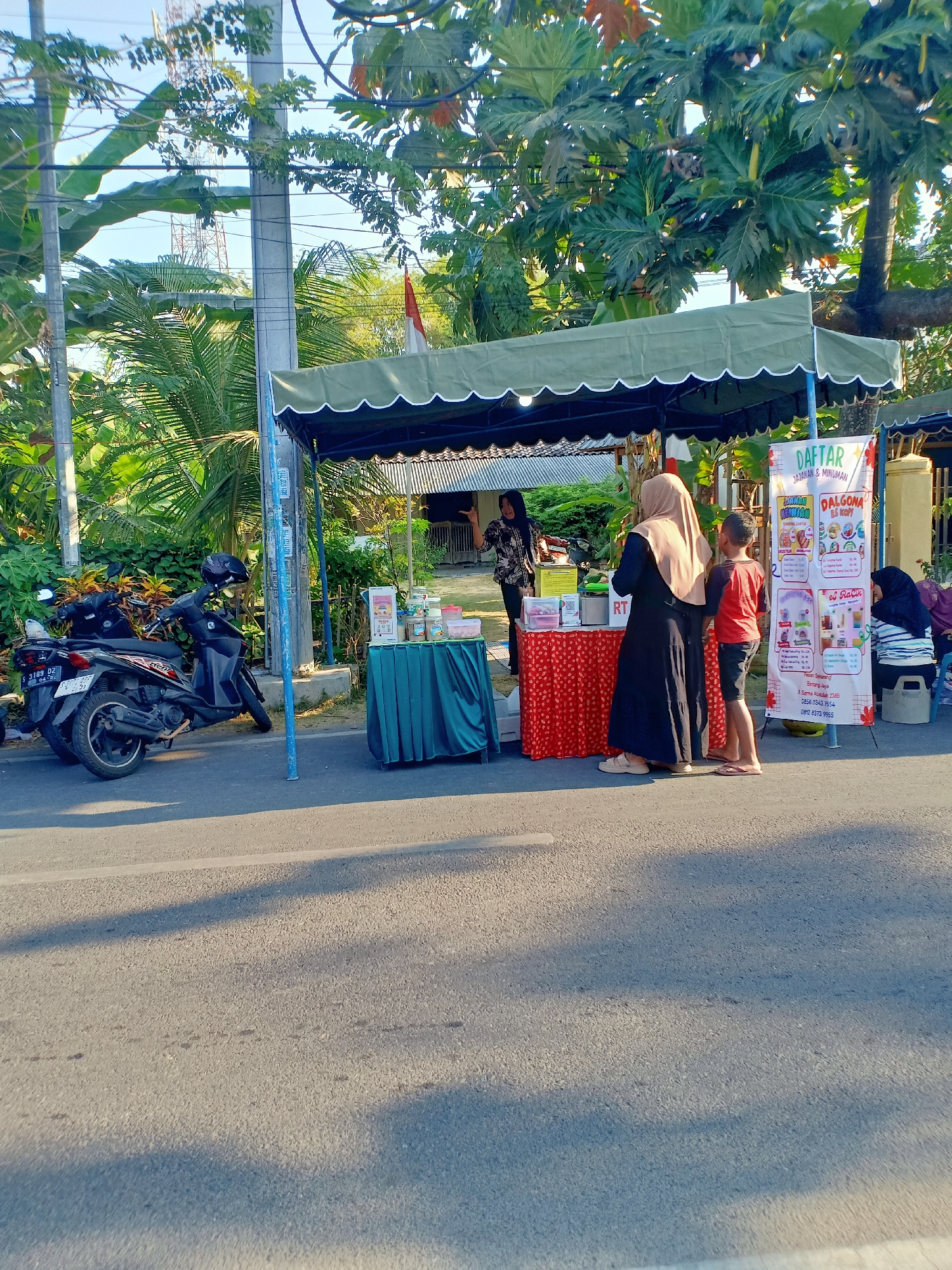 The image depicts a street-side food stall in Bojonegoro, Indonesia, bathed in the warm light of late afternoon. A green tent is set up to provide shade, with various food and drink items displayed on tables covered with red and green tablecloths. A banner, written in Indonesian, advertises "DAFTAR JAJANAN & MINUMAN" (List of Snacks and Drinks) and lists items like "ES KOPI DALGONA" (Dalgona Coffee Ice) and "ES RACIK".
Several people are present at the stall. Two women and a young boy are standing in front of a table, appearing to interact with the vendors. One woman, wearing a hijab, is looking at the items on display. Another woman, dressed in a patterned blouse, seems to be attending to customers. A third woman, also wearing a hijab, is seated to the right of the stall, with a child nearby. Two motorbikes are parked on the side of the road next to the stall, suggesting a common mode of transportation in the area.
The environment is a typical Indonesian street scene, with lush green vegetation, including palm trees and larger leafy trees, surrounding the area. Power lines and a utility pole are visible, indicating an urban or semi-urban setting. The road surface is asphalt. The overall atmosphere is one of a bustling local market or roadside eatery.