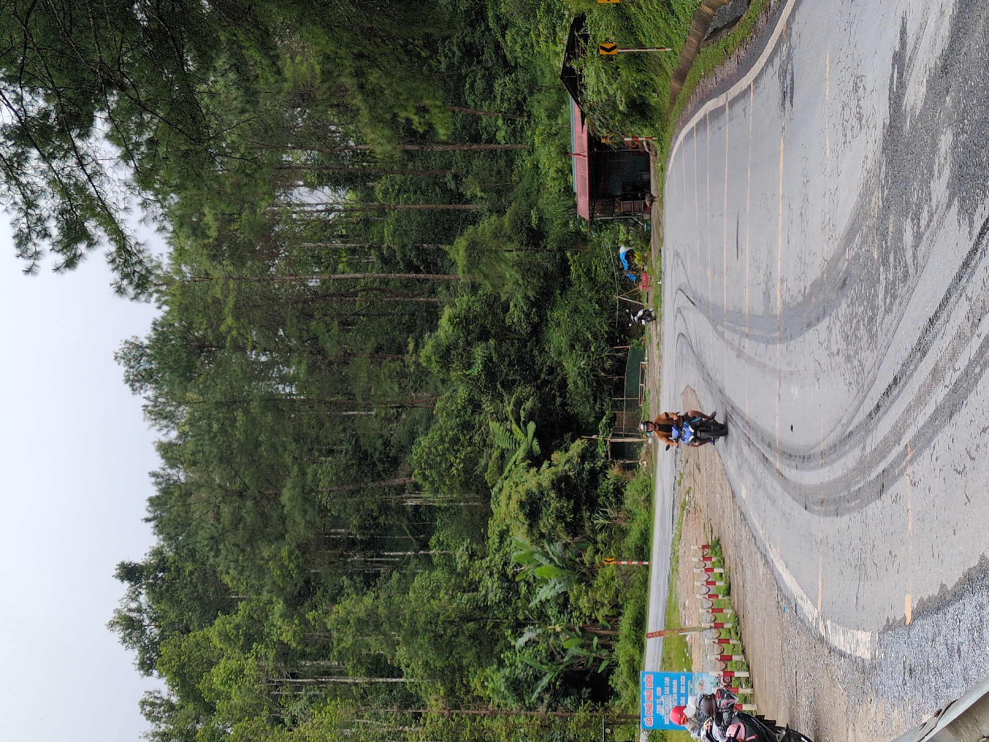 A motorcyclist is riding on a wet, asphalt road that curves to the right in a forested area. The road has tire tracks and yellow lane markings. The surrounding environment is dense with green trees and vegetation, with a clear sky overhead. To the right of the road, there is a small, rustic building with a red roof. A yellow directional sign is visible on the right side of the road. In the distance, other motorcycles and a sign with Vietnamese text are present near the edge of the road on the left. The overall scene depicts a rural road in Quản Bạ Commune, Vietnam.