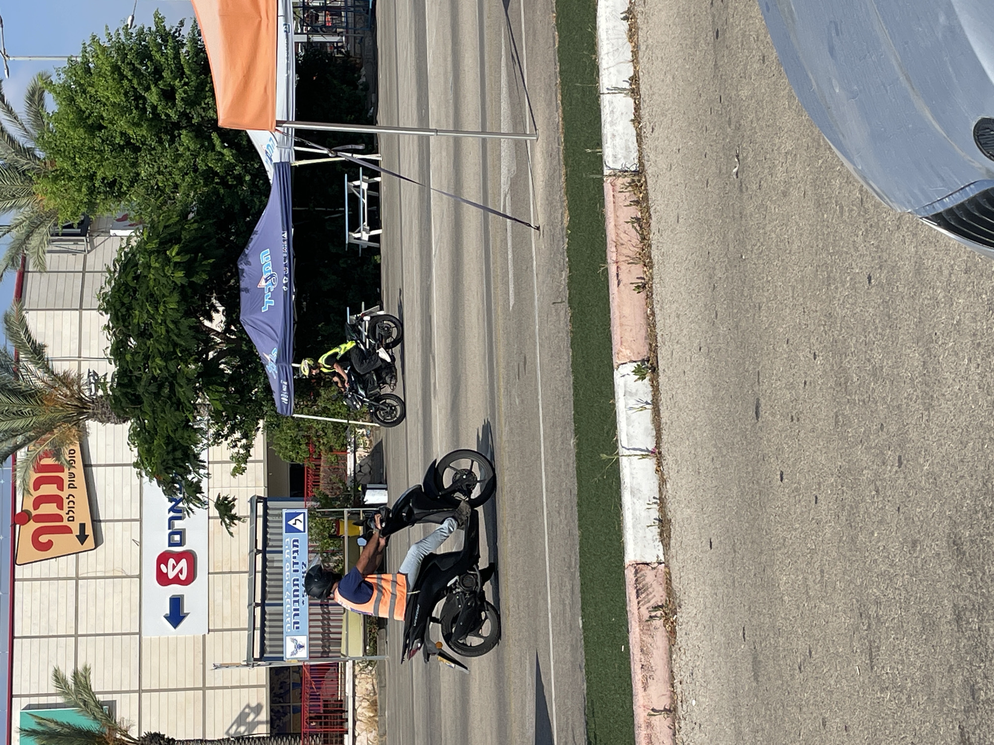 This image depicts a street scene in Haifa, Israel, likely in the vicinity of a commercial area. In the foreground, a person wearing a reflective orange vest and a helmet is riding a black scooter. The scooter has a visible brand name "VISION" on its side. The rider is wearing blue jeans.
In the background, a building with a facade of beige tiles is visible. Above this building, a large red sign with Hebrew text appears to be an advertisement or store name. Adjacent to this, a blue arrow points to a red symbol within a white square, also featuring Hebrew text.
Further back, two tents are set up. The one on the left is blue with Hebrew lettering, and the one on the right is orange. Under the blue tent, a picnic table is visible, and a person in a yellow safety vest and helmet is seated on a motorcycle. Another motorcycle is parked near the blue tent.
The environment appears to be a sunny day, with palm trees and green foliage contributing to a semi-urban setting. The road surface is asphalt, with painted lane markings. A concrete curb with pink and white markings separates the road from a grassy area. A portion of a silver car is visible in the bottom right corner of the image.