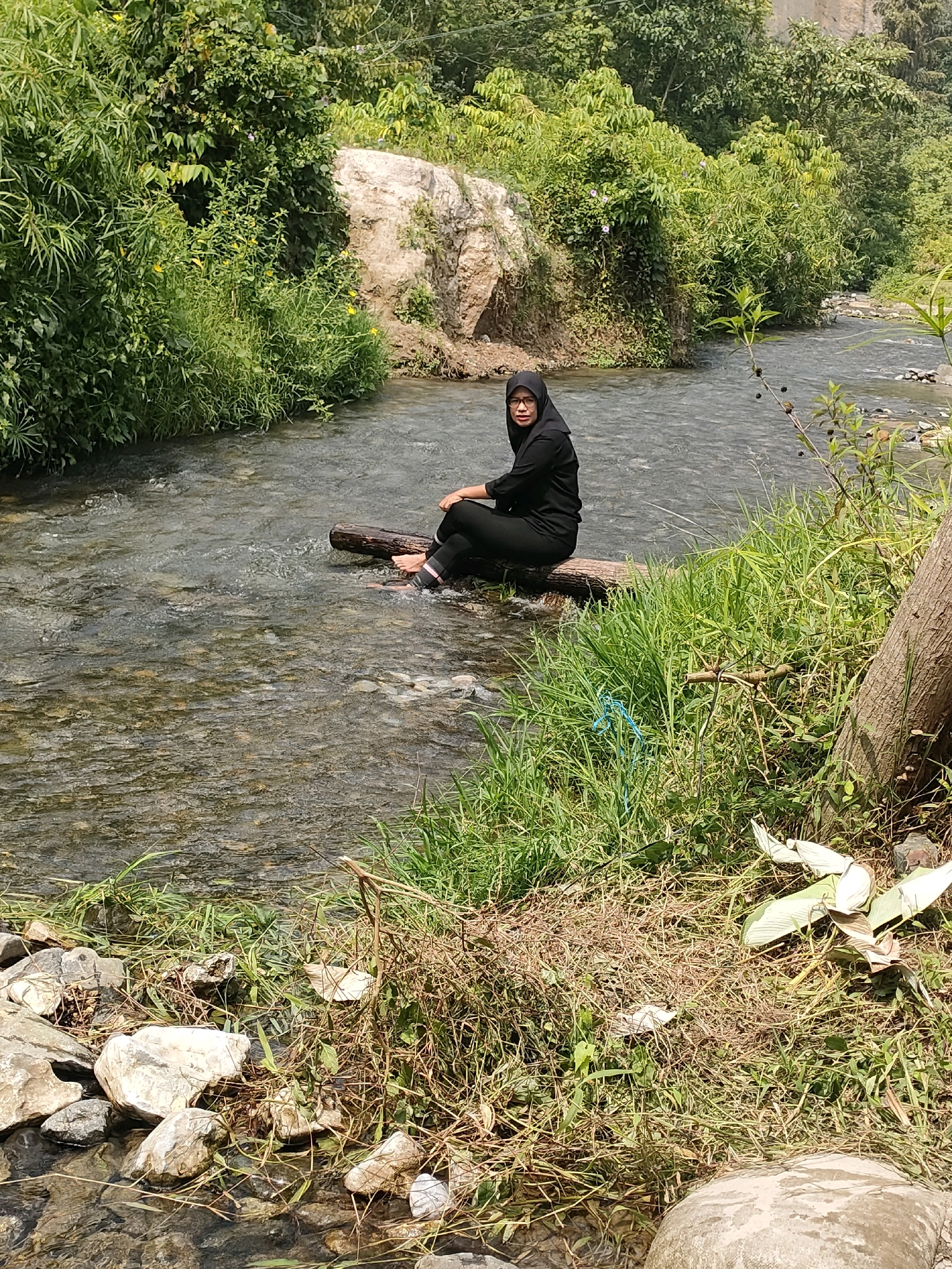 In the city of Sianok VI Suku, Indonesia, a woman wearing a black hijab and black clothing is seated on a log that is partially submerged in a shallow, clear river. Her feet are in the water. The riverbank is covered in lush green grass and vegetation, with a large rock formation visible in the background. Trees and dense foliage line the river, creating a natural, serene environment. The scene is captured in daylight under a clear sky.
