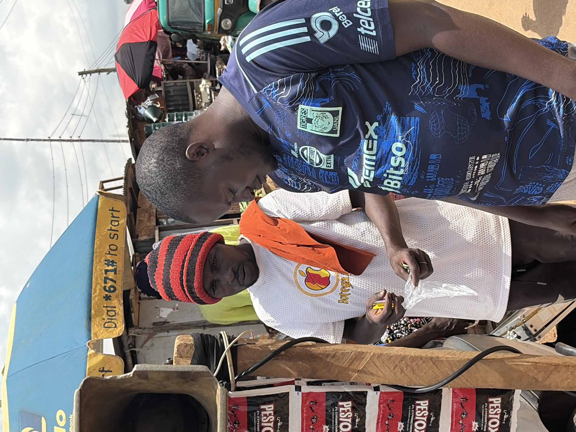 In Gura Top, Nigeria, two men stand in front of a market stall. The man on the left wears a red and black striped beanie and a white t-shirt with an apple logo and the word "Botega." He holds yellow packets and a clear plastic bag. The man on the right wears a dark blue t-shirt with various logos, including "Tigres," "CEMEX," and "Bitso." An orange cloth is draped over the shoulder of the man on the left. Behind them, a blue and yellow umbrella with the text "Dial *671# to start" is visible, along with other market stalls, umbrellas, and a green motorized tricycle. Rolls of what appear to be pest control products are stacked vertically on the left side of the image. The ground is dirt.