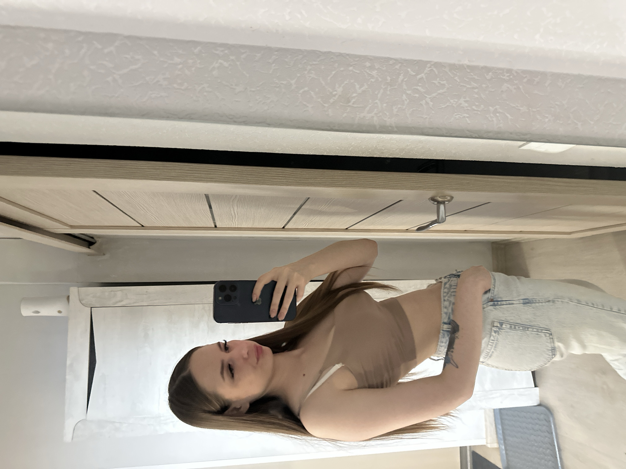 A woman is taking a selfie in a room of unknown location. The environment includes walls, a door, and a vanity with a mirror. She is wearing a nude-colored top and light blue jeans. She is holding a smartphone in her left hand to take the photo.