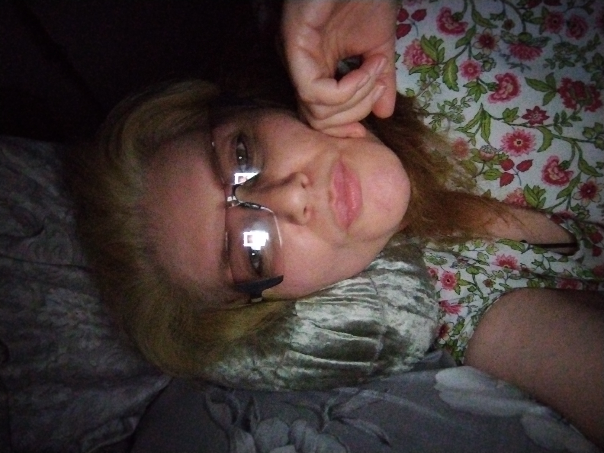 An adult female subject, wearing eyeglasses and a floral top, is depicted in a close-up portrait, oriented sideways. Her head rests on a textured, silvery-grey pillow, with her light-colored hair fanning out. Her gaze is directed towards the camera, with bright, rectangular reflections, consistent with a digital screen, visible in her eyeglass lenses. Her lips are slightly parted. One hand, with fingers lightly curled, rests near her temple. The surrounding environment includes floral-patterned fabric (her top) and dark, subtly patterned bedding. The scene is located in Al Mas'oudi, United Arab Emirates.