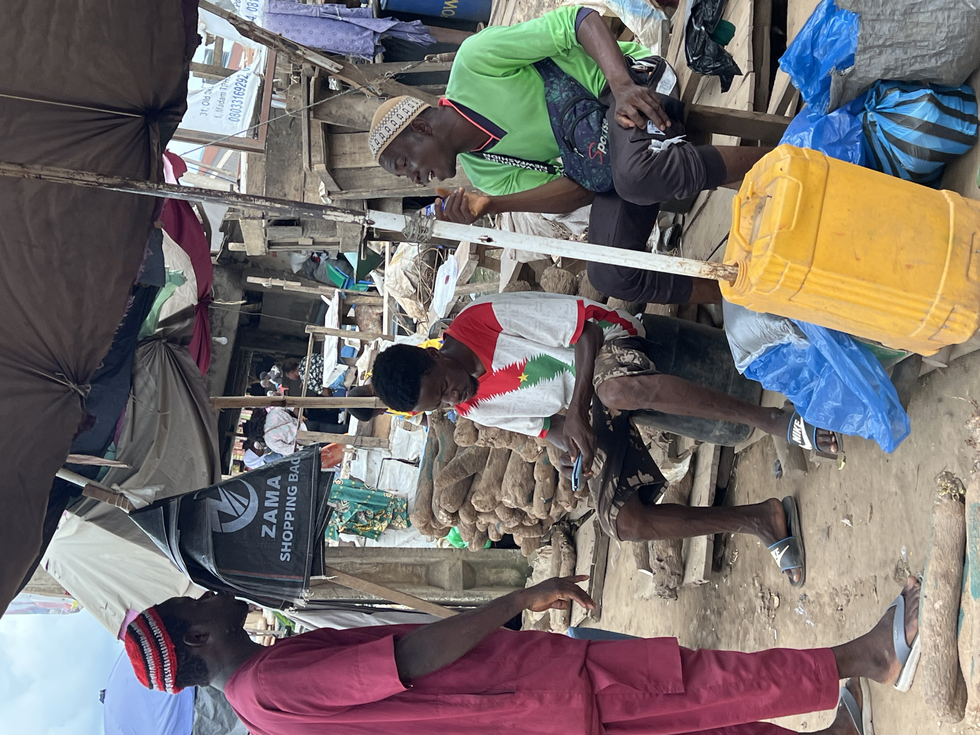A medium shot captures three men in a Nigerian market, likely in Asaba, Nigeria, based on visual cues consistent with such a setting. The man on the left, wearing a maroon robe and a red and white striped cap, stands facing the center, with his left arm down and his right arm bent at the elbow. He appears to be in mid-conversation. In the center, a man with a red, white, and green t-shirt and camouflage shorts sits on a black drum, engrossed in his mobile phone. To his right, another man in a green v-neck shirt and dark pants sits on a wooden bench. He wears a patterned cap and holds a small object in his right hand while speaking, gesturing with his left hand.
The environment is a bustling market with numerous stalls and vendors. Overhead, a large, weathered tarpaulin provides shade. In the background, stacks of large, yam-like tubers are visible, suggesting a food market. Various market goods, including bags and plastic containers, are scattered around, with a prominent bright yellow jerrycan in the foreground. The ground is dusty and uneven. The overall scene depicts a typical day of commerce and social interaction in an open-air Nigerian market.