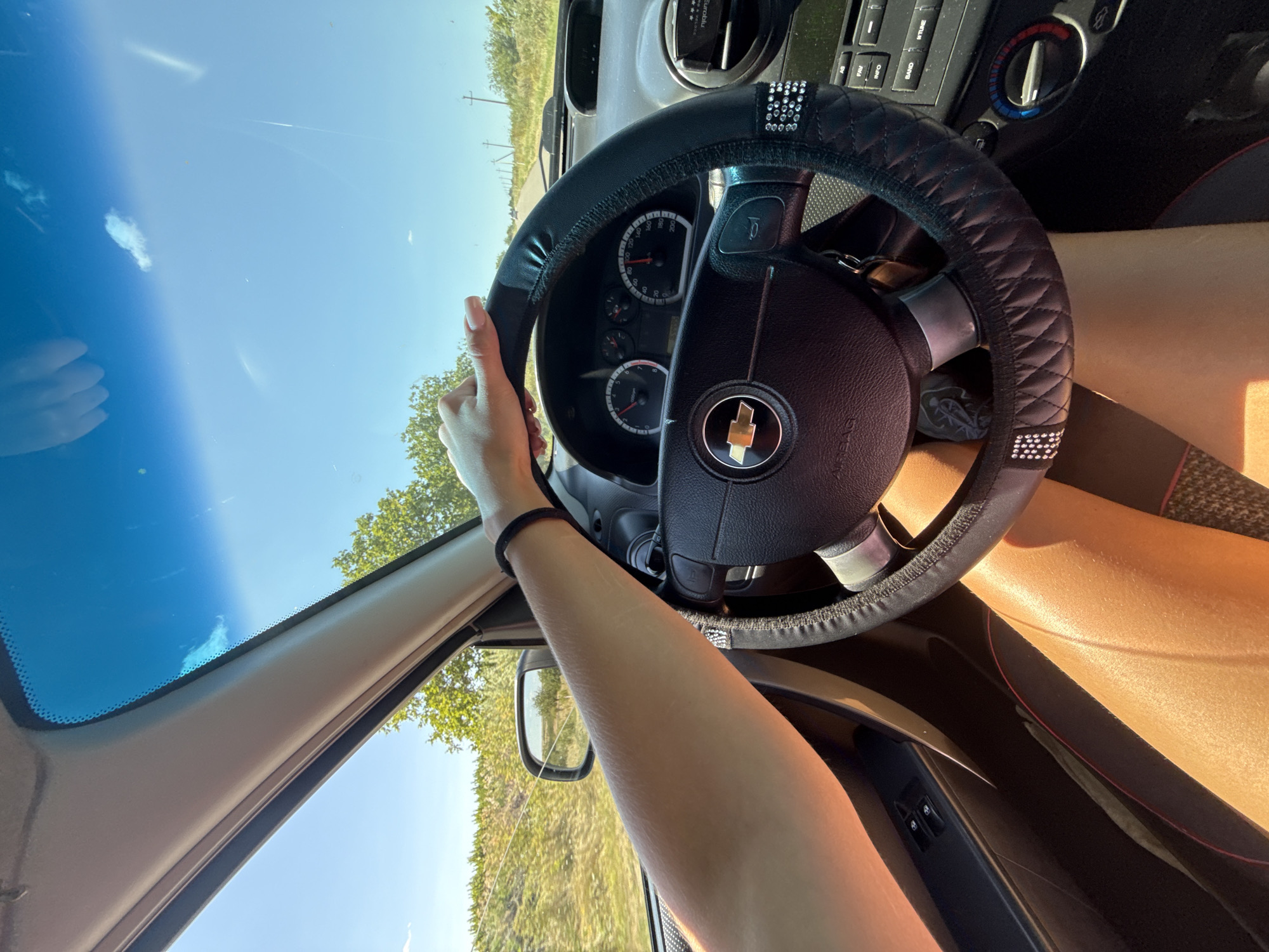 The image shows a first-person perspective from inside a car, looking forward. A person's arm and hand are visible, resting on the steering wheel. The steering wheel is covered with a black quilted material and adorned with rhinestones. The dashboard features a speedometer and other gauges. Outside the car, a rural landscape with trees and a road can be seen under a clear blue sky. The scene appears to be captured in Basarabeasca, Moldova, suggesting a drive through the countryside.