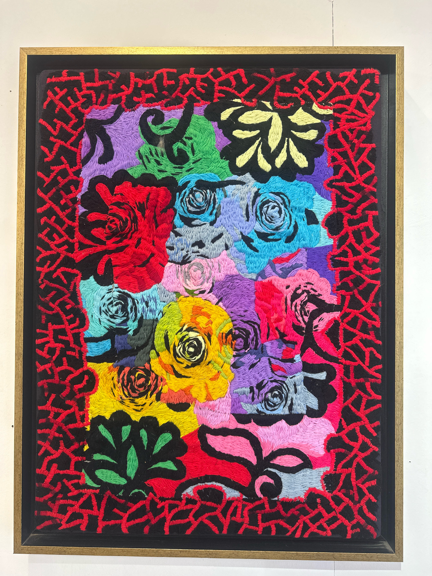 An embroidered artwork featuring a dense arrangement of stylized floral elements in vibrant, saturated colors is displayed in a gold-toned frame. The artwork is hanging on a white wall, presumably in a gallery or exhibition space, located in the city of Mate Asher Regional Council, Israel. The artwork has a black backing with a decorative border composed of red stitching that creates a complex, interwoven pattern along the perimeter of the main image. The central subject matter consists of various flowers, primarily roses, depicted in a Fauvist or Expressionistic style. The colors used include vivid reds, blues, purples, greens, yellows, pinks, and black. The textures are created by the visible stitches of the embroidery, giving the artwork a rich, tactile quality. The flowers are densely packed, with some overlapping and intermingling, creating a sense of depth and visual complexity. The overall impression is one of bold color and dynamic composition. No people or specific environmental details beyond the exhibition setting are visible in the image.