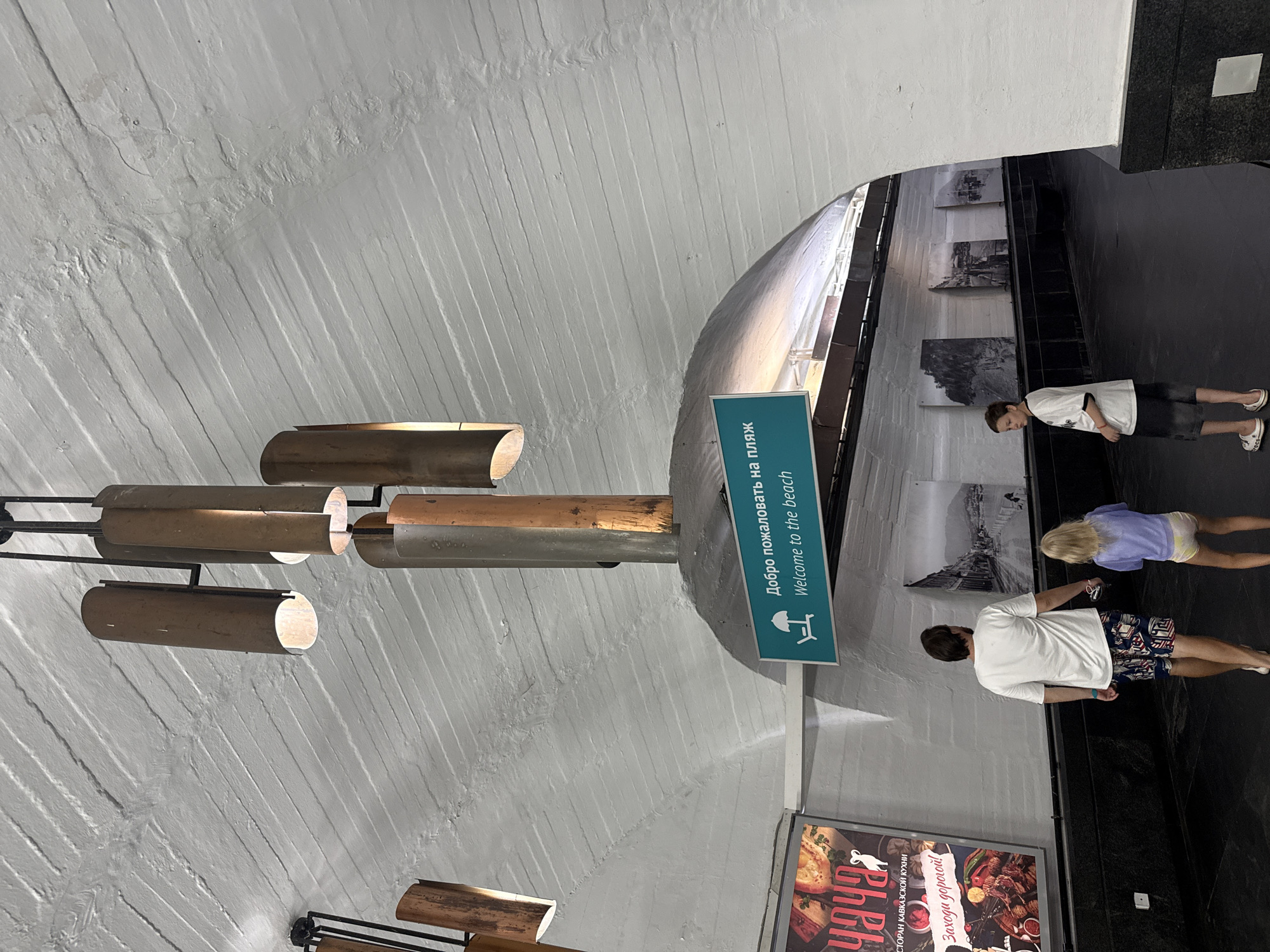 In a curved, tunnel-like passageway with a white, textured ceiling, a family consisting of a man and two children walks away from the camera towards a sign that reads "Добро пожаловать на пляж Welcome to the beach" with a graphic of a beach chair and umbrella. The man wears a white t-shirt and patterned shorts. The younger child, a girl, wears a light purple t-shirt and light-colored shorts. The older child, a boy, wears a black and white t-shirt and dark shorts. The passageway features black tiled walls and is adorned with large black and white photographs of coastal scenery. On the left, a colorful advertisement for a restaurant called "УЧА" is visible, showcasing food items. The lighting appears to be artificial, emanating from overhead fixtures that are cylindrical and metallic in appearance. This scene appears to be a pedestrian underpass or tunnel within the city of Yalta, Ukraine, likely leading towards a beach area, given the prominent signage.