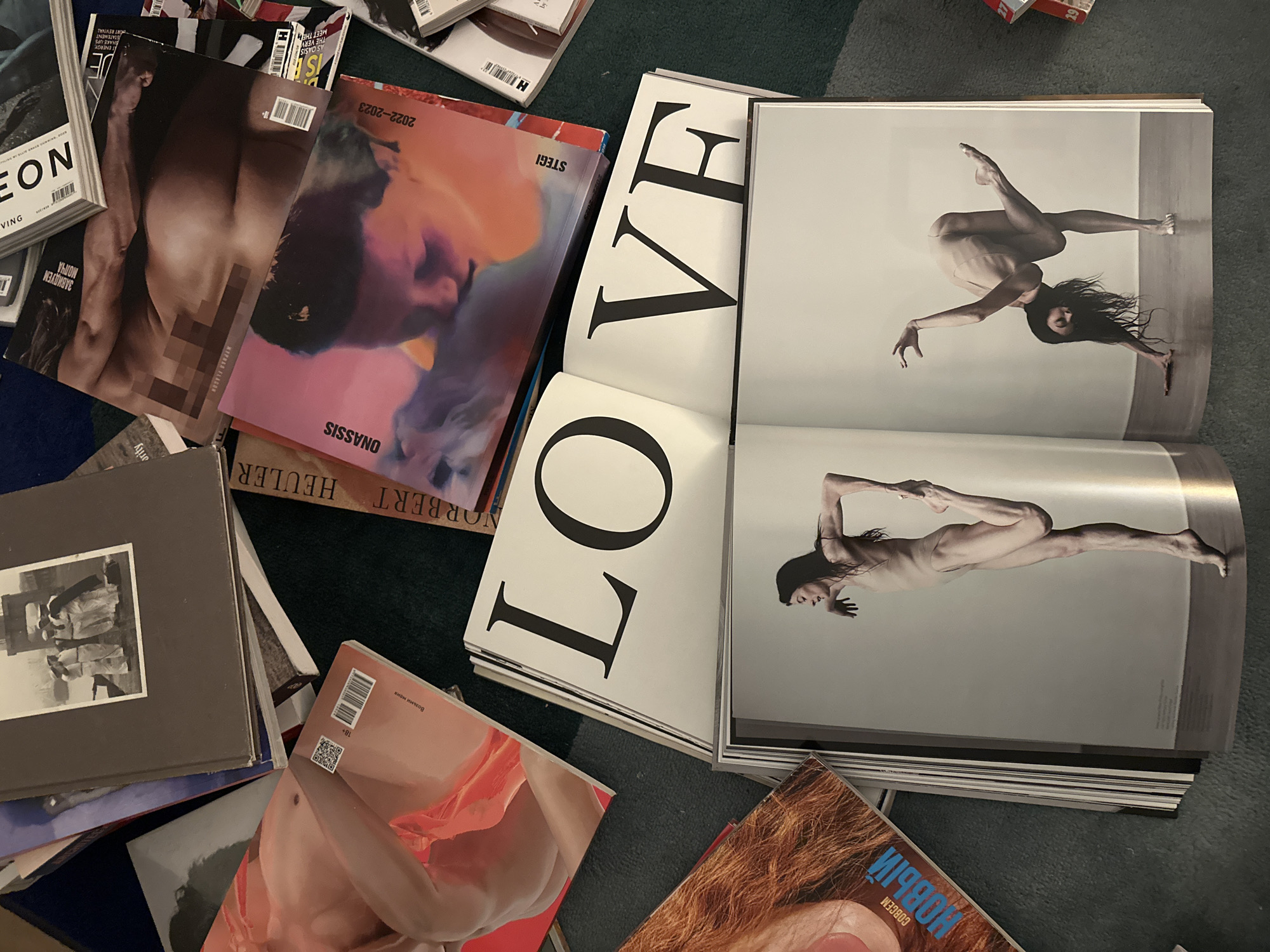 The image shows a pile of magazines and books scattered on a dark green or blue carpet. Prominently featured is an open magazine titled "LOVE" with large black lettering. Inside this magazine are two full-page, grayscale images of a slender woman in athletic poses, possibly dance or yoga. Other magazines are stacked around this central book, including one titled "ONASSIS" and another titled "STEGI 2022-2023." A magazine titled "MIPRAS FLACON" is also visible, partially obscured by other items. There is also a magazine cover featuring a blurred image of a male torso and another titled "LEON." The overall arrangement suggests a casual, possibly disorganized, collection of print media. The image does not contain specific information to identify a location in London, United Kingdom.