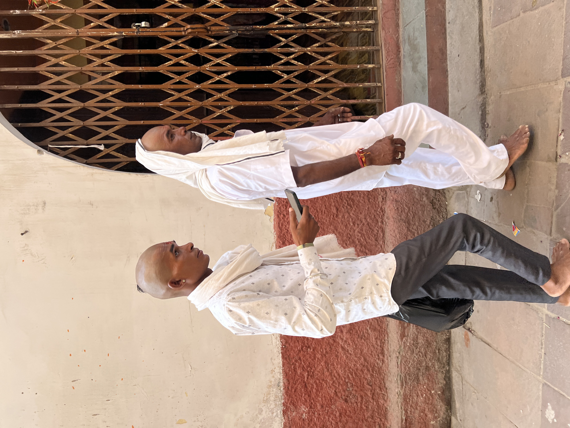 Two men are walking past a building with a large, intricate metal gate in Pushkar, India. The man on the left is wearing a white patterned shirt, dark pants, and a white scarf around his neck. He has a shaved head with a small tuft of hair at the back and is holding a mobile phone. He is also carrying a black bag. The man on the right is wearing a white outfit, including loose-fitting pants and a short-sleeved shirt, and has a white cloth draped over his head. Both men are barefoot. The building behind them has a textured reddish-brown base and a cream-colored upper section. The metal gate is ornately designed with diamond-shaped openings. The ground is paved with stone tiles.