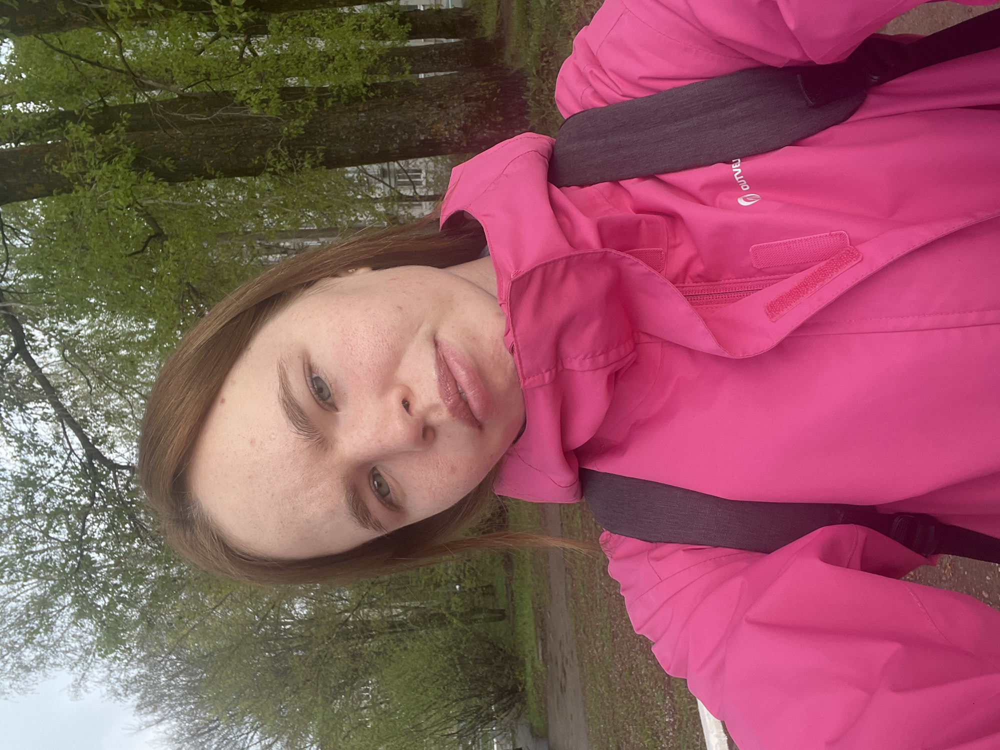 A selfie of a Caucasian female wearing a pink jacket and a backpack is taken outdoors. The jacket has a hood and visible Velcro closures. The backpack straps are dark gray. The woman has light brown hair and freckles on her face. The background shows a park or wooded area with trees bearing green leaves. The image is oriented vertically. The precise location within the city of Chaikovsky, Russia is not deducible from the given visual information.
