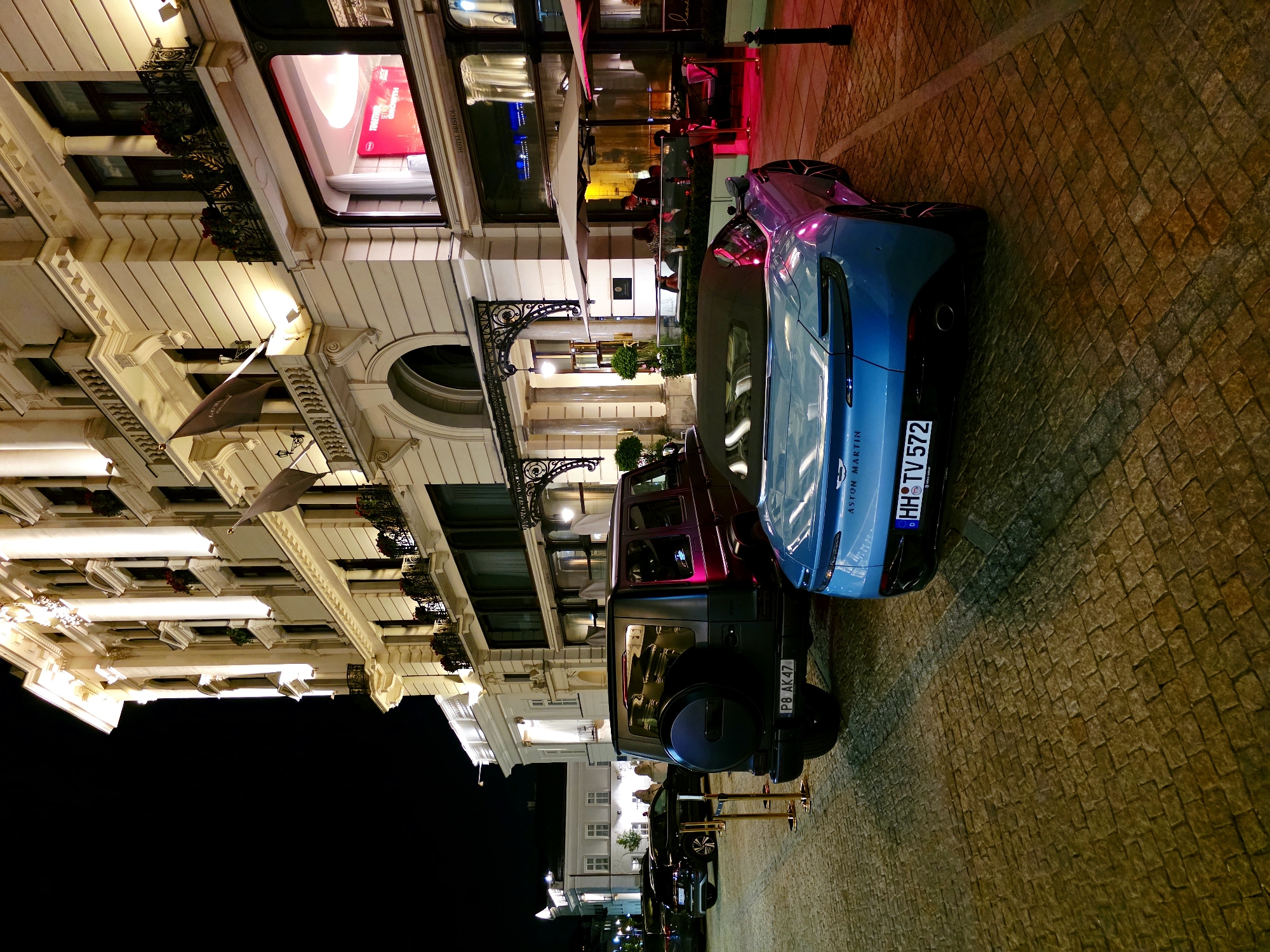 This is a nighttime exterior shot of the Hotel Bristol in Warsaw, Poland, which is located at Krakowskie Przedmieście 42/44. The hotel is a grand, cream-colored building with ornate architectural details, illuminated by warm exterior lighting. In the foreground, two luxury cars are parked on a cobblestone street. To the left is a dark-colored SUV, possibly a Mercedes-Benz G-Class, with a spare tire mounted on the back and a license plate that reads "PB AK47". To the right is a bright blue convertible sports car, which appears to be an Aston Martin, with its license plate reading "HH TV 572". The Aston Martin has its top down and is angled towards the viewer. Beyond the cars, the hotel features a prominent entrance with large columns and decorative wrought iron gates. A restaurant or cafe area with outdoor seating is visible to the right, under large umbrellas, with diners seated inside and illuminated by pinkish lighting. The sky is dark, indicating it is nighttime.