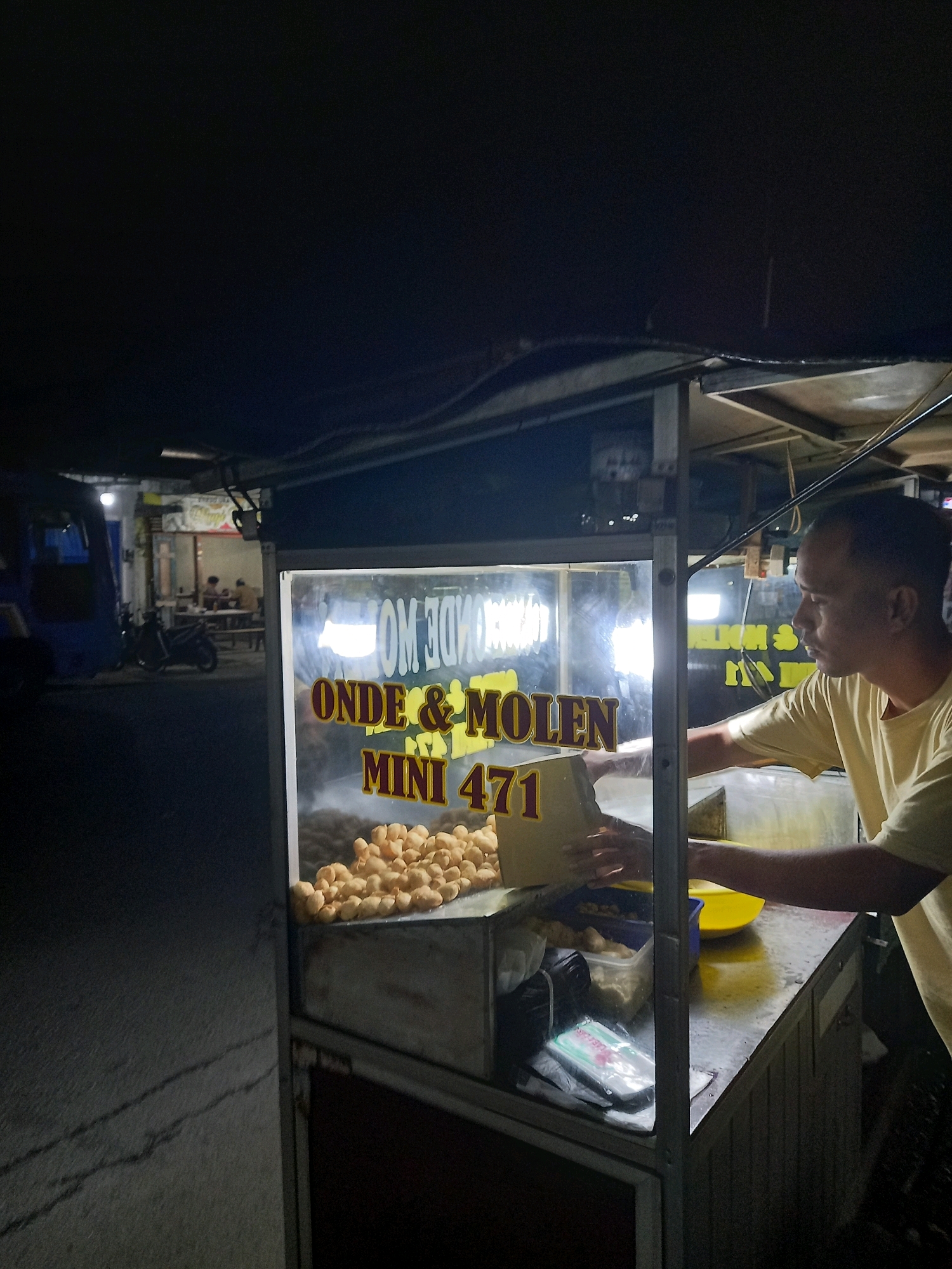 The image depicts an outdoor food stall at night, likely in the city of Bawang, Indonesia. A man wearing a yellow t-shirt is standing behind a glass display counter filled with fried snacks. Text on the glass reads "ONDE & MOLEN MINI 471". The stall has a metal frame and is illuminated by artificial lighting, creating reflections on the glass. In the background, a blue vehicle and a motorcycle are visible, along with a few people seated at tables in what appears to be a dimly lit street or outdoor seating area. The overall atmosphere suggests a typical street food vendor setting.