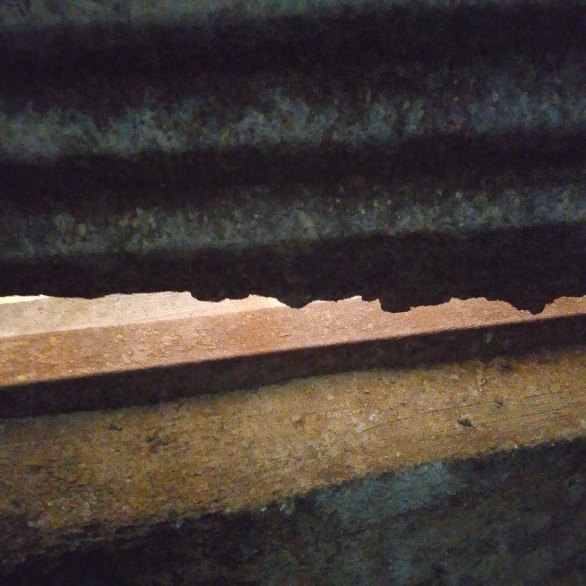 The image shows a close-up view of weathered, corrugated metal and wooden planks, likely part of an outdoor structure. The metal appears rusty and has a dark, textured surface with horizontal ridges. Below the metal, there are two wooden planks. The upper plank is reddish-brown and shows signs of wear, with peeling paint or a similar surface degradation. The lower plank is a lighter, more yellowish-brown color and also appears aged and rough. A gap is visible between the metal and the upper plank, allowing a sliver of light to penetrate. The scene suggests an industrial or rustic setting, possibly an abandoned or utilitarian structure. There is no indication of the specific location within Jalingo, Nigeria, as the image focuses solely on the materials and textures.