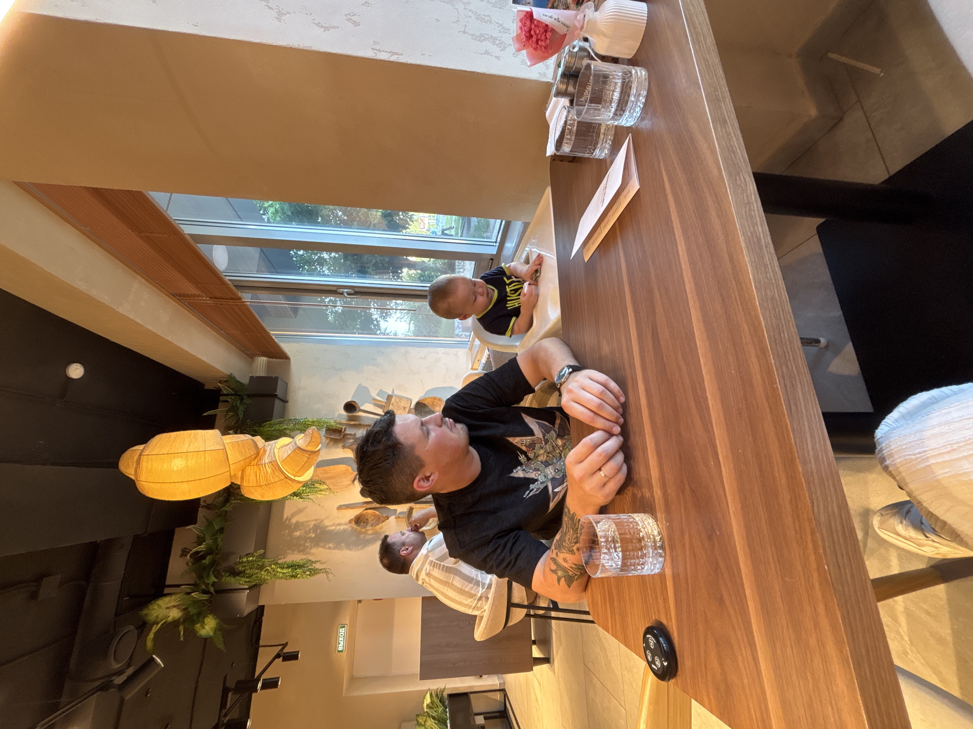 In a well-lit restaurant with warm-toned walls and a black ceiling, a man with tattoos on his arms sits at a wooden table, looking to his right. He is wearing a black t-shirt. In front of him are two glasses of water, a pager, and what appears to be a menu or bill. To his right, a baby sits in a high chair and looks down at the table. In the background, another man in a striped shirt is seated at a table, facing away from the camera, and further back, a sign indicating an "Exit" in Russian (ВЫХОД) is visible. Large windows with blinds look out onto a green, outdoor area. A decorative lamp hangs from the ceiling, and plants are visible in the background. The overall scene suggests a casual dining experience.