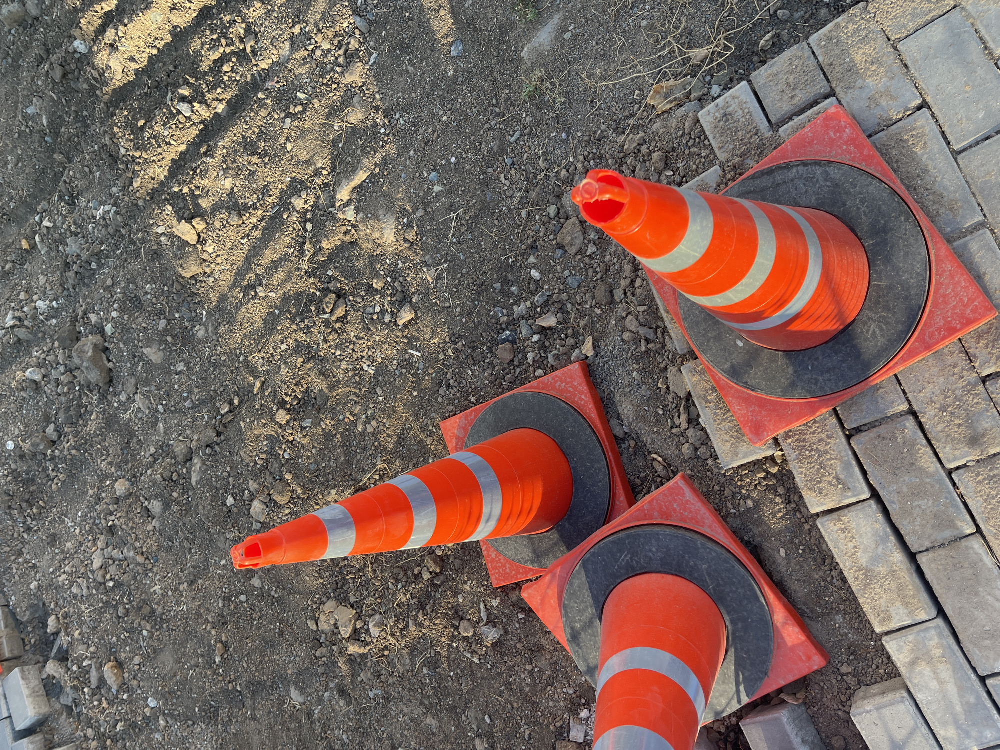 The image shows three orange traffic cones with reflective silver bands. Two of the cones are placed on a surface of grey rectangular paving stones, and the third cone is positioned on a dirt and gravel surface to the left of the paved area. The cones appear to be set up as a temporary barrier or to mark a hazard.
The scene is lit by sunlight, casting distinct shadows from what appear to be tire tracks or some uneven ground on the dirt surface. This suggests it is daytime, likely morning or late afternoon, given the angle of the shadows. The weather appears to be dry.
The setting suggests an outdoor construction site or area undergoing repair. The presence of both dirt/gravel and paving stones indicates a transition zone or an area with mixed ground surfaces. There is no visible text in the image. The overall impression is one of a functional, work-in-progress environment.