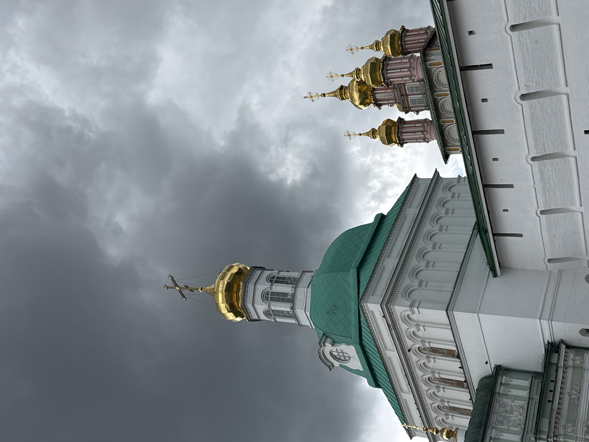 This is a photograph of the Trinity Lavra of St. Sergius in Sergiev Posad, Russia, under a dramatic, cloudy sky. The image features several religious structures, including a prominent white building with a large green domed roof, topped with a smaller golden dome and a cross. To the right, a group of smaller, golden domes, also topped with crosses, rise against the dark clouds. A white wall with decorative cutouts runs across the foreground, partially obscuring the lower portions of the buildings. There are no people visible in the frame. The overall impression is one of imposing architecture set against a moody, overcast sky.