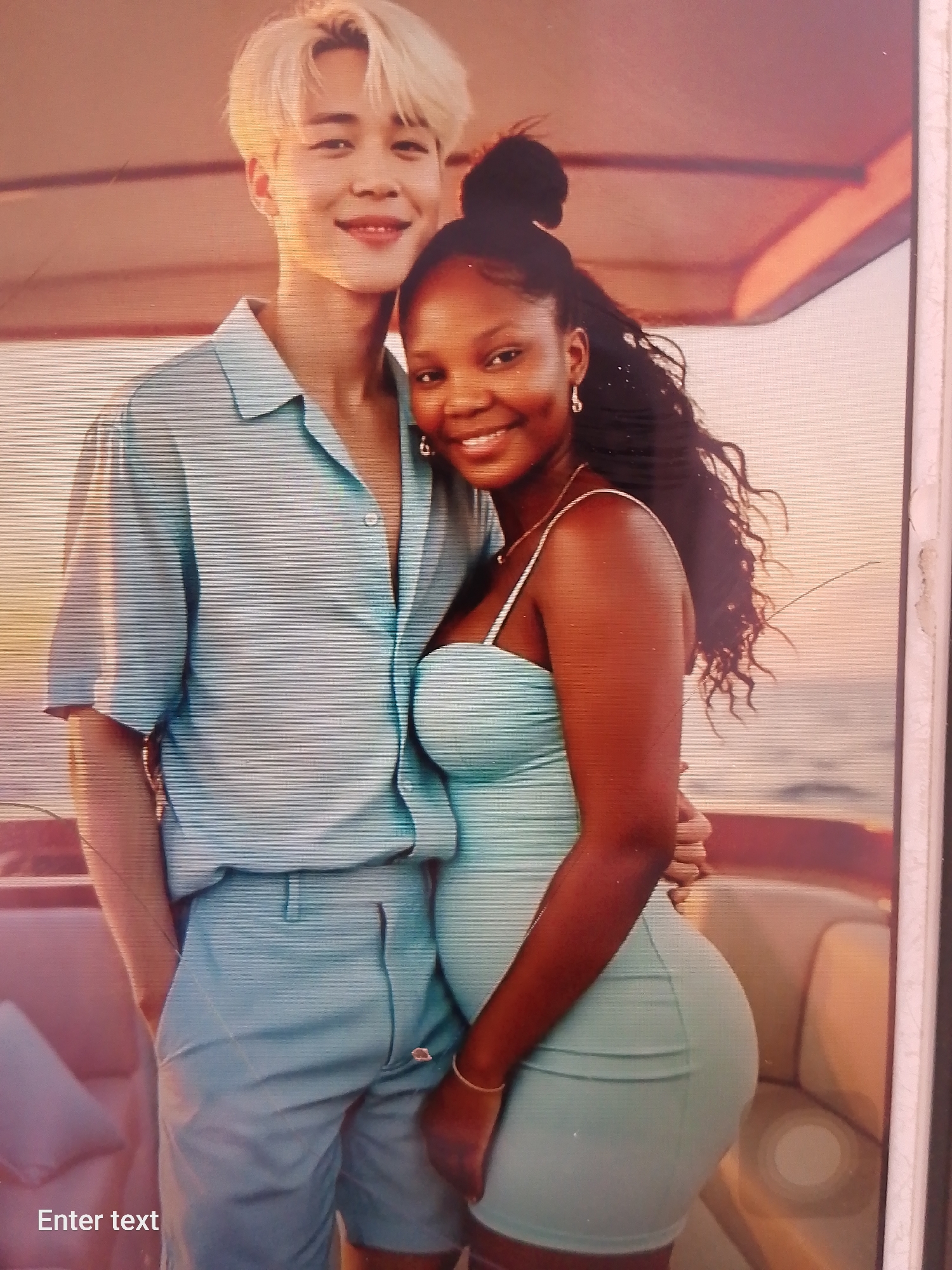 A photograph shows a woman with dark skin, curly hair pulled into a bun, and a light green dress, posed next to a man with light skin, blond hair, and a matching light green shirt and shorts. They are standing close together with the woman's arm around the man's waist, both smiling at the camera. The background suggests a boat, with a visible ocean and sunset behind them. No indication of Adankolo, Nigeria, can be seen in the image.