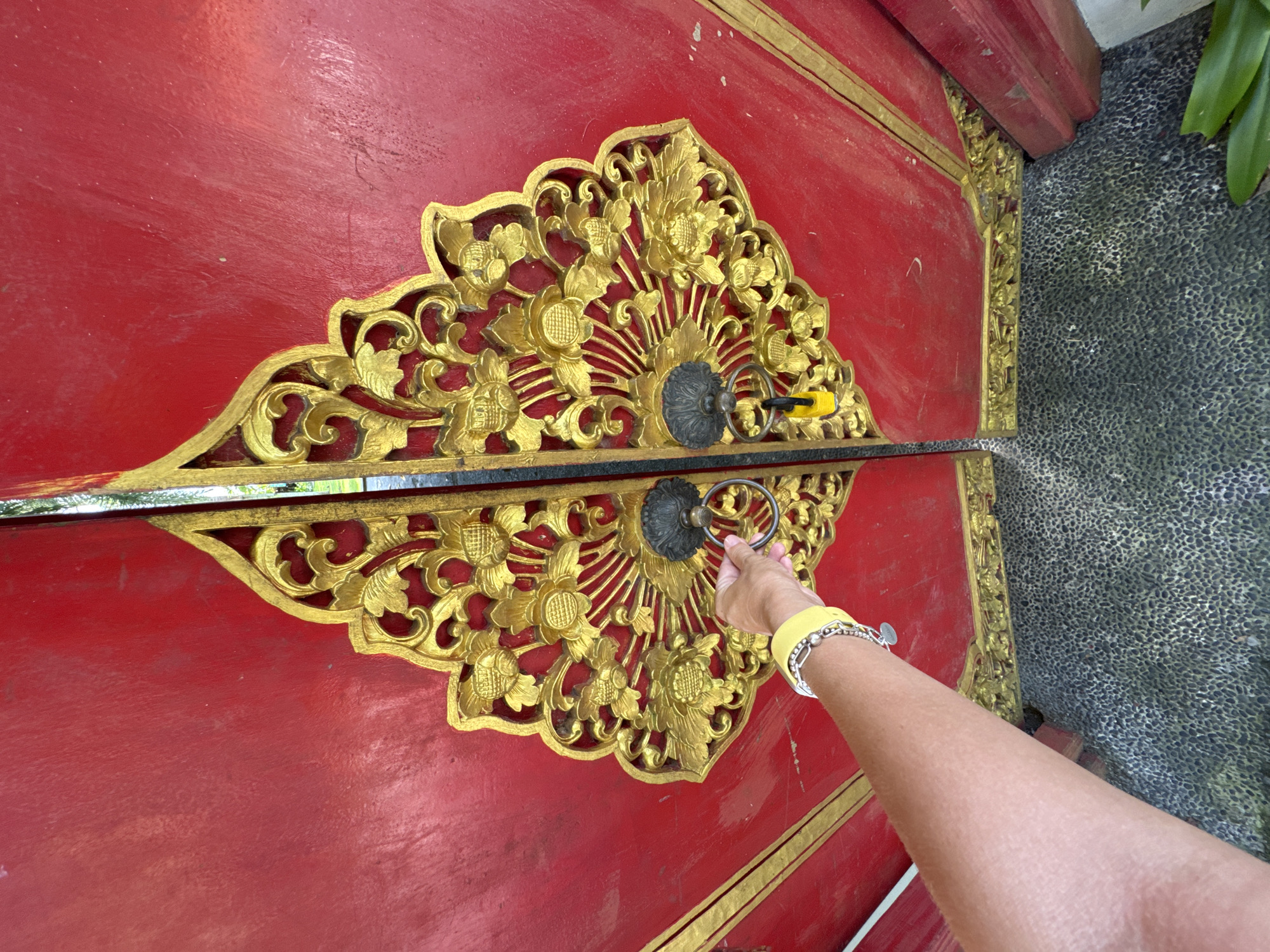 A person's arm is reaching toward a decorated door in Kuta Selatan, Indonesia. The door is painted red with ornate gold carvings that meet in the middle, featuring circular knobs with rings for handles. The person is wearing a yellow watch and silver bracelet on their wrist. The door is set against a textured, dark gray wall.