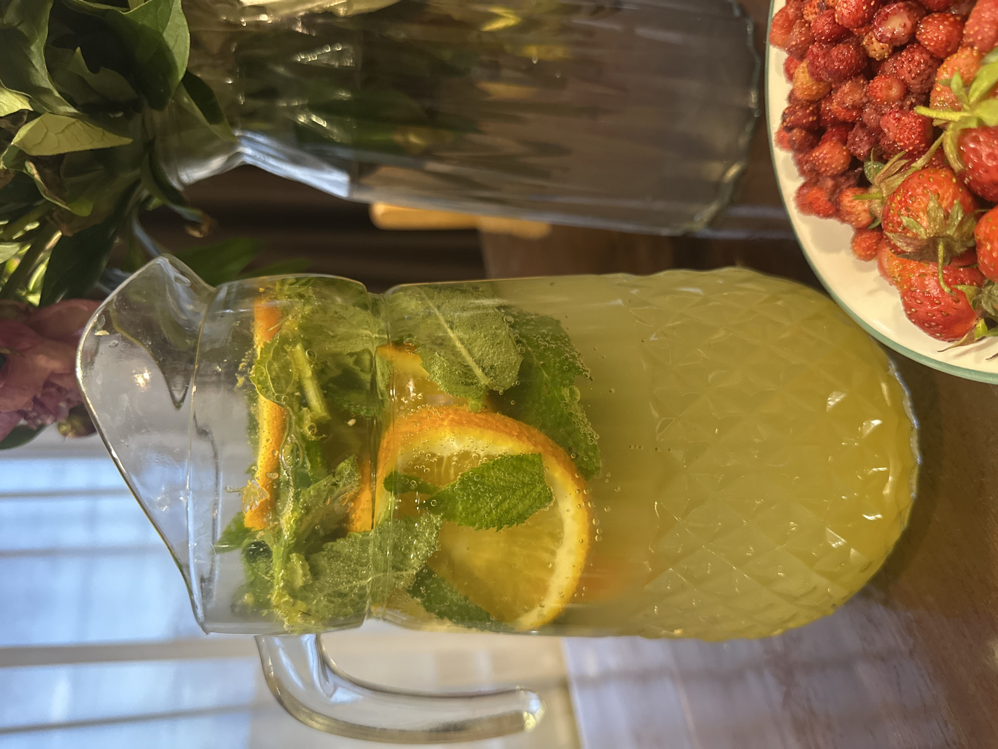 This media file displays a close-up, angled shot of a pitcher filled with a yellow, bubbly beverage, likely lemonade or a similar citrus drink. Slices of orange and fresh mint leaves are visible submerged in the liquid, adding to its refreshing appearance. The pitcher itself is made of clear glass with a diamond-patterned texture on its base. Bubbles are present throughout the drink, suggesting it is carbonated or has just been mixed.
To the right of the pitcher, a white plate is filled with a pile of small, red wild strawberries. Some larger strawberries are also visible among them. The background includes a glimpse of a bouquet of green leaves and what appears to be a pink flower to the left, and a blurred, light-colored surface to the lower left, possibly a table. The overall setting suggests a warm, perhaps summery, outdoor or indoor gathering where refreshments are being served. The lighting is soft, indicating it could be daytime. There is no visible text in the image. The scene evokes a sense of refreshment and enjoyment.