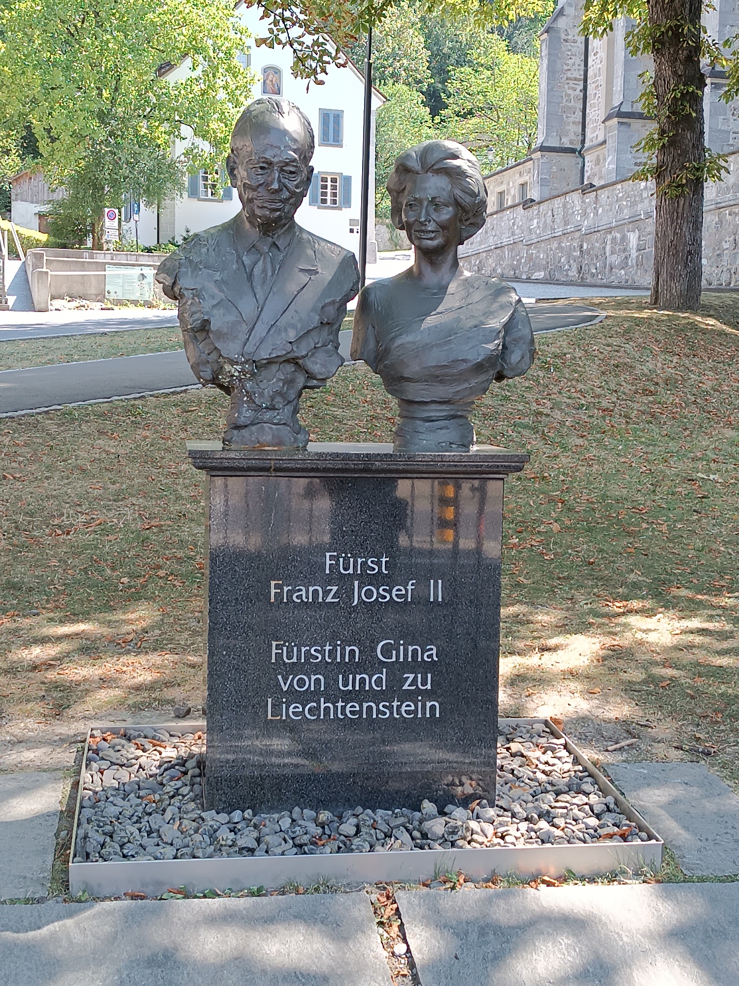 A bronze bust of a man and a woman, identified by an inscription as Fürst Franz Josef II and Fürstin Gina von und zu Liechtenstein, stands on a dark granite pedestal in a park-like setting. The man's bust, on the left, depicts him wearing a suit and tie, with his head turned slightly to his right. The woman's bust, on the right, shows her with her head tilted back and to her left, her hair styled in an updo. The pedestal has the inscription "Fürst Franz Josef II" on the top line, and "Fürstin Gina von und zu Liechtenstein" on the second line. The busts are positioned in front of a low stone wall, with a paved walkway and grass in the foreground. Trees with green foliage provide shade. Behind the wall, a building with light-colored walls and blue shutters is visible. The scene is illuminated by sunlight, casting shadows on the ground. This memorial is located in Vaduz, Liechtenstein, near the Prince's Palace.