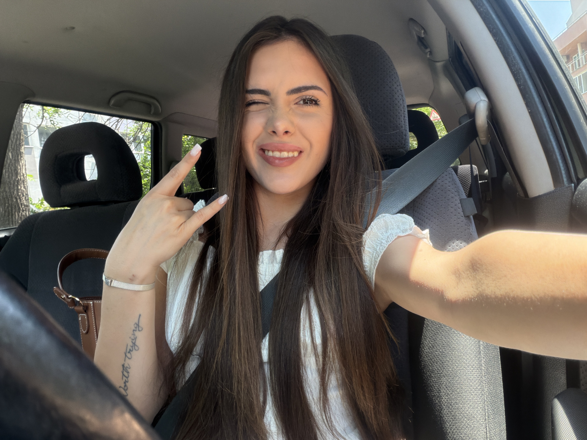The image is a selfie taken by a young woman from inside a car, with the setting indicated as Burgas, Bulgaria. The woman, appearing in her 20s, has long, straight dark brown hair, and is seen smiling broadly while winking with her left eye. She makes a playful "rock on" or "devil horns" hand gesture with her left hand, which features light-colored, possibly white, manicured nails. A silver-colored bracelet adorns her left wrist, and a script tattoo that reads "worth trying" is visible on her left forearm.
She is wearing a light-colored top with textured or ruffled details on the sleeves and has a seatbelt fastened across her chest. The car's interior features dark fabric seats and headrests, with a glimpse of the steering wheel on the left. Bright daylight is evident, suggesting a sunny day, with trees, green foliage, and a building visible through the car windows in the background. A small, brown top-handle handbag is also partially visible on the left side, behind her arm. The woman's expression conveys happiness and playfulness.
