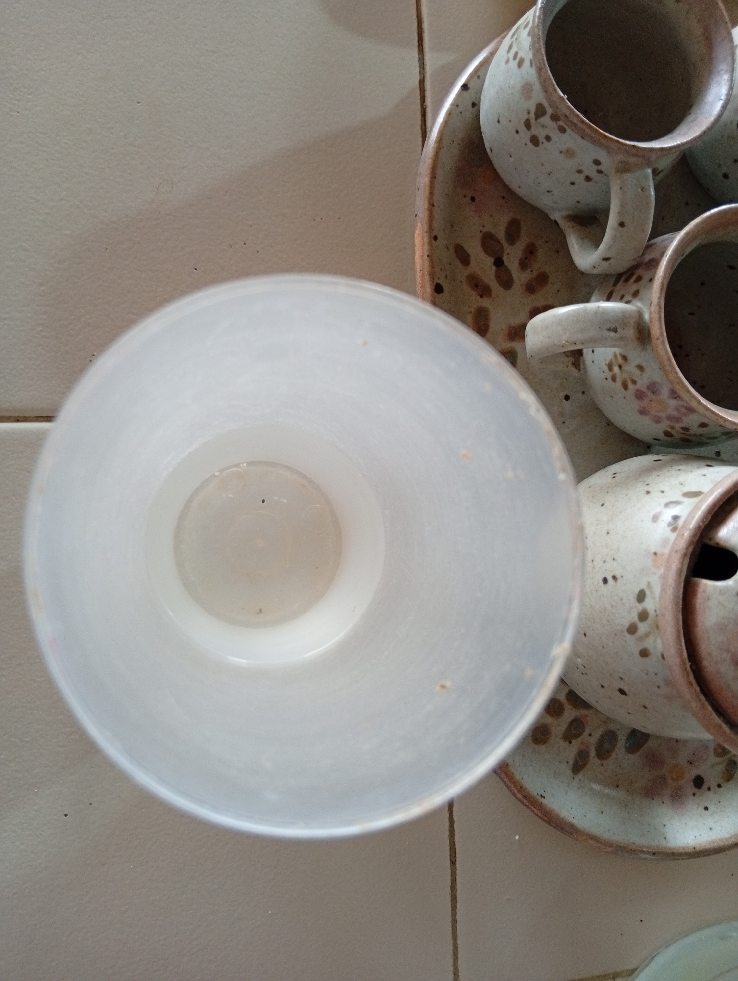 A close-up, overhead shot captures a collection of rustic, speckled ceramic mugs and saucers arranged on a tiled surface. In the foreground, a large, translucent white cylindrical container is positioned, obscuring part of the scene. Behind it, several mugs and saucers are visible. One mug, partially in focus, sits on a saucer, its handle facing upwards and to the right. Another mug, more in focus, is placed further back, also on a saucer, with its handle oriented similarly. A third mug is partially visible behind the first. The ceramic pieces feature a light-colored base with irregular dark brown speckles and some scattered floral designs in pink and yellow. The tiled surface beneath appears to be a light beige color with visible grout lines. The overall impression is of a still life arrangement in a domestic setting, possibly a kitchen or dining area in San Antonio de Los Altos, Venezuela.