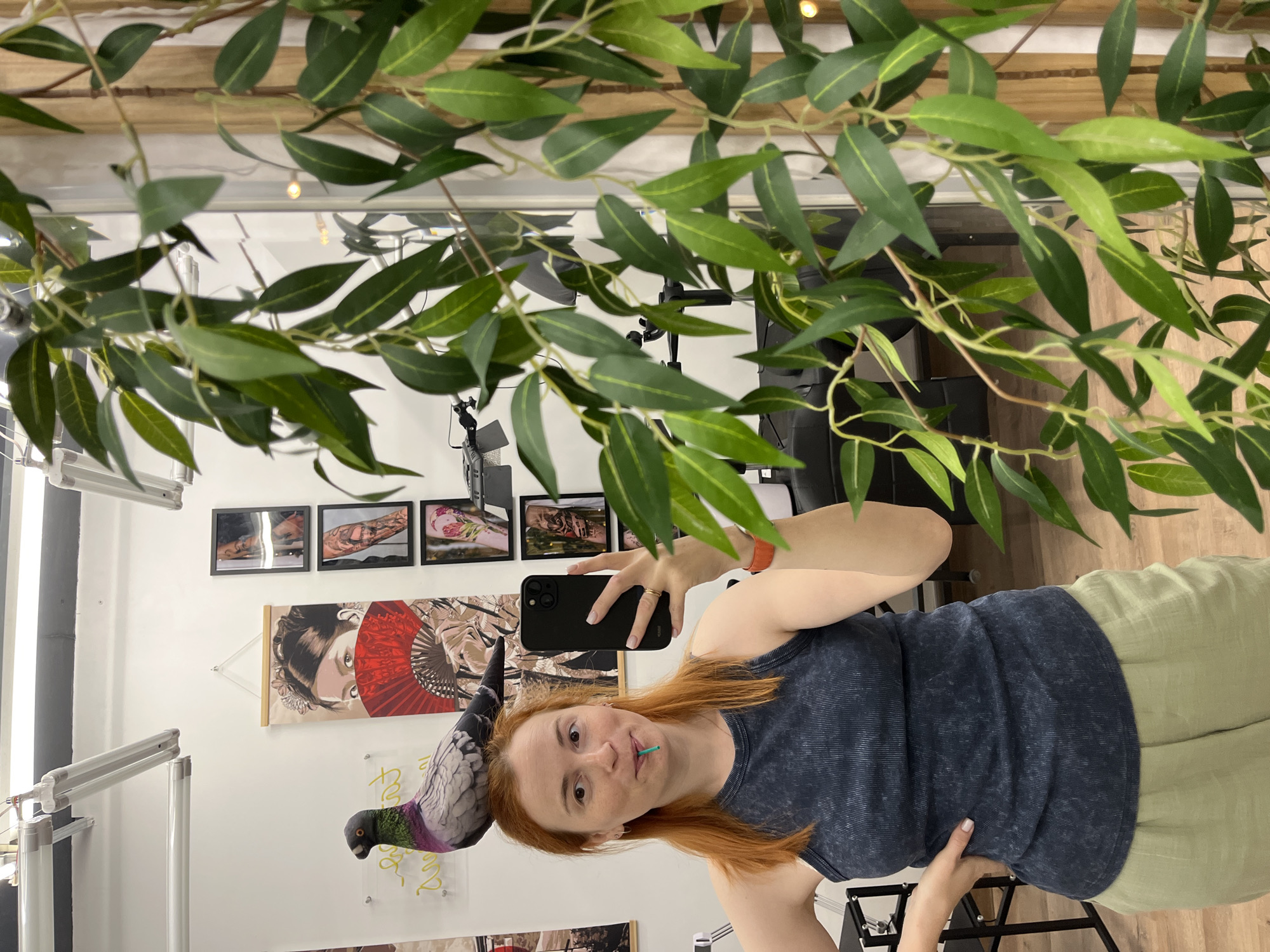 A woman with auburn hair and a pigeon perched on her head stands in a room that appears to be a tattoo studio. She is wearing a dark blue ribbed tank top and light green linen pants. In her right hand, she holds a black smartphone, and a green straw is placed in her mouth. The pigeon on her head is a typical city pigeon with grey, black, and iridescent purple and green plumage. Behind her, the wall is decorated with several framed tattoo flash art prints and a long banner depicting a woman holding a red fan. A large artificial plant with green leaves is visible in the foreground to the right. The scene is brightly lit, likely by artificial lighting visible on the ceiling.
