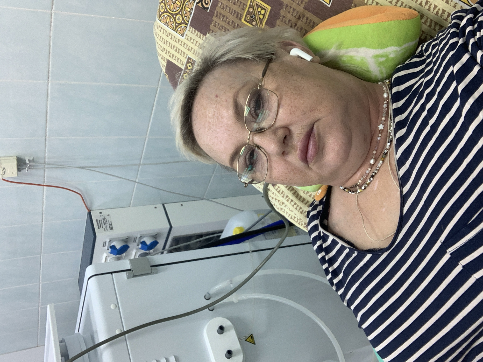 The image depicts an older Caucasian woman with short blonde hair and glasses lying on a patterned pillow with a green and orange cushion behind her head. She is wearing a striped blue and white shirt and several necklaces. An earbud is visible in her right ear. To her left is a medical device, possibly a dialysis machine, with tubes and connections. The background consists of light blue tiled walls. Based on the available information, the location is the city of Chaikovsky, Russia.