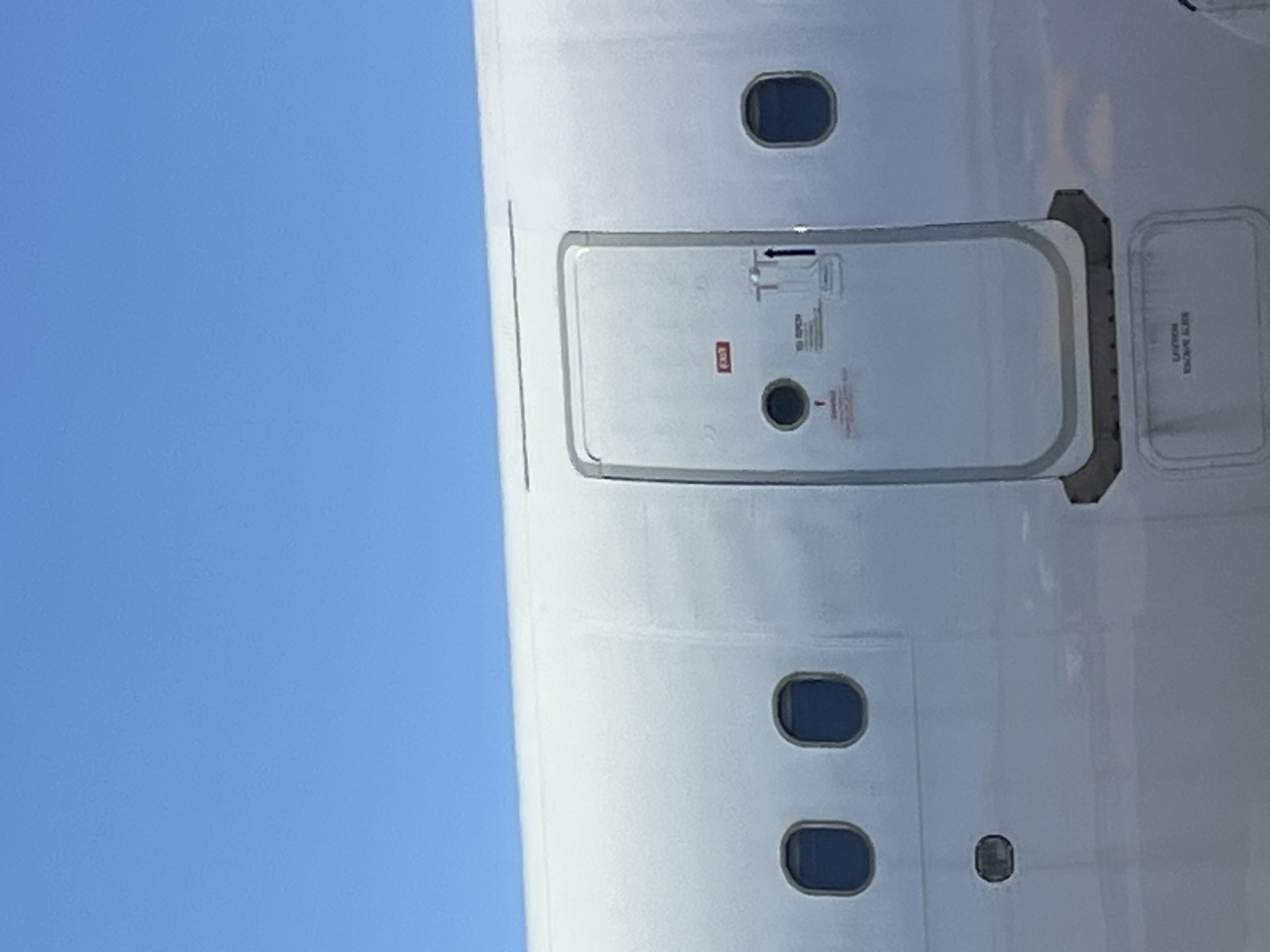 The image shows the exterior of an airplane, likely at Cam Ranh Airport in Cam Ranh Bay, Vietnam. The fuselage is white, with several oval-shaped windows visible. A prominent exit door, also white with grey trim, is open. Red signage indicating "EXIT" is visible on the door, along with instructions on how to open it, including an upward arrow and the text "TO OPEN". Below the open door, a rectangular panel with text indicating "CAUTION" and "ESCAPE SLIDE" is present. The sky above is a clear, bright blue. The scene is static and appears to be a daylight shot.
