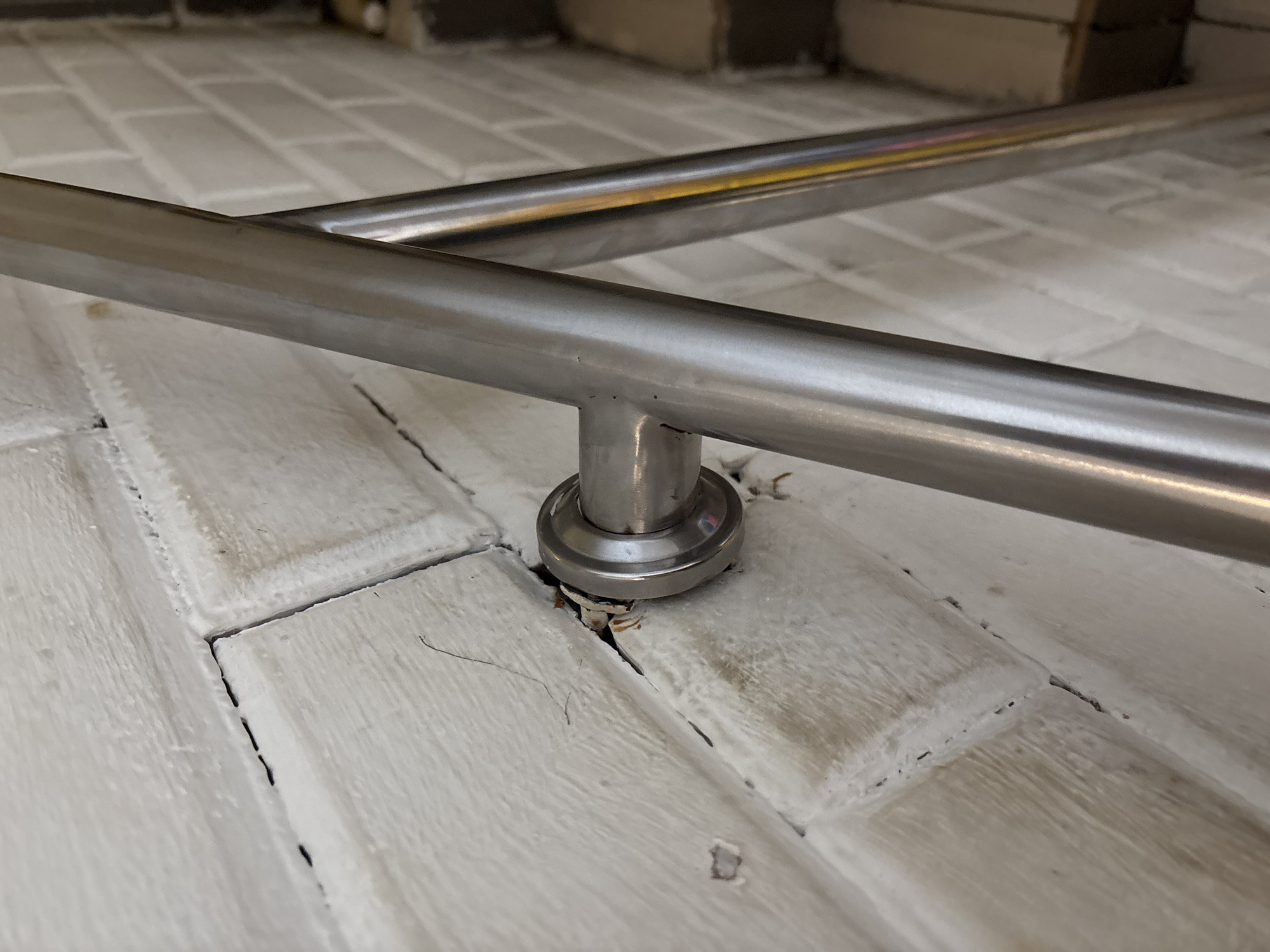 A close-up shot shows a silver metal handrail attached to a white brick wall. The handrail is secured to the wall by a circular bracket. The brick wall appears to be aged, with visible cracks and some areas of chipped paint. The mortar between the bricks is dark, creating a contrast with the white brick. In the background, a portion of stairs is visible, also appearing to be made of brick and painted white. The lighting suggests an indoor environment. The scene depicts a functional element of a building's architecture, specifically a staircase railing. This scene is located on the third floor of the Pekanbaru Grand Mosque, Pekanbaru City, Riau Province, Indonesia.