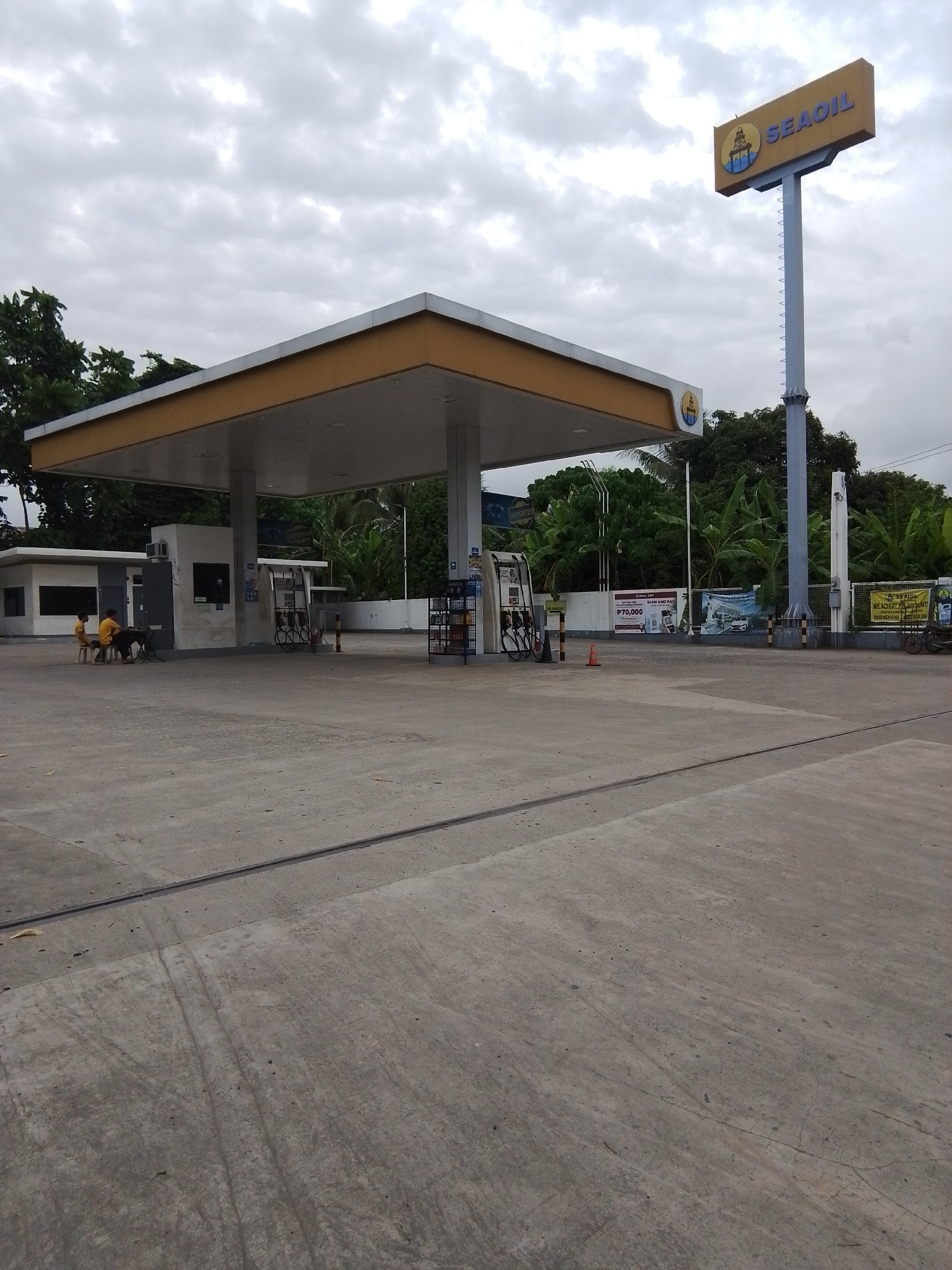 A wide shot captures a SEAOIL gas station under a cloudy sky in Davao City, Philippines. A prominent yellow and blue SEAOIL sign stands tall on a pole to the right. The gas station canopy, also yellow and white, covers multiple gas pumps. Two individuals, wearing yellow shirts, are seated on stools near a building on the left, with a dark-colored dog beside them. Various advertisements are visible on the walls behind the gas pumps. The ground is paved with concrete, showing tire marks and some debris. Lush green trees and vegetation form the background.