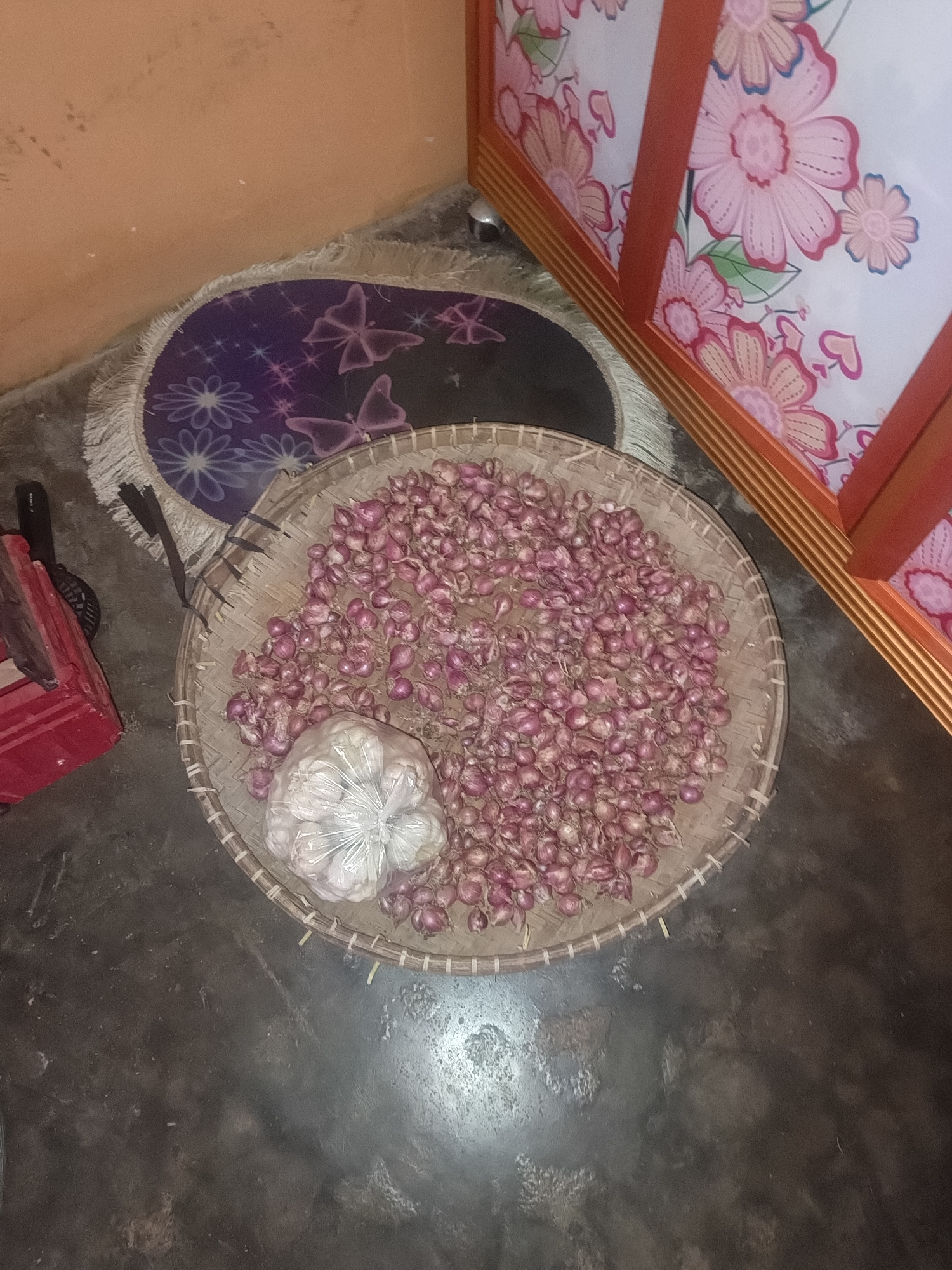 The image shows a close-up, high-angle view of a woven basket filled with small, pink onions. To the left of the basket is a bunch of garlic wrapped in clear plastic. The basket sits on a dark, rough-textured floor. In the background, to the left, a portion of an orange wall is visible above a round rug with a purple and black pattern featuring butterflies and flowers. To the right of the basket, a cabinet with a red frame and white doors decorated with floral patterns is visible. The scene appears to be indoors, possibly in a kitchen or storage area. There are no people or animals visible in the image. The lighting suggests an indoor setting with artificial light sources. The overall impression is one of domesticity and food preparation or storage. It is not possible to determine the exact location within Terbanggi Marga, Indonesia, from the provided image.