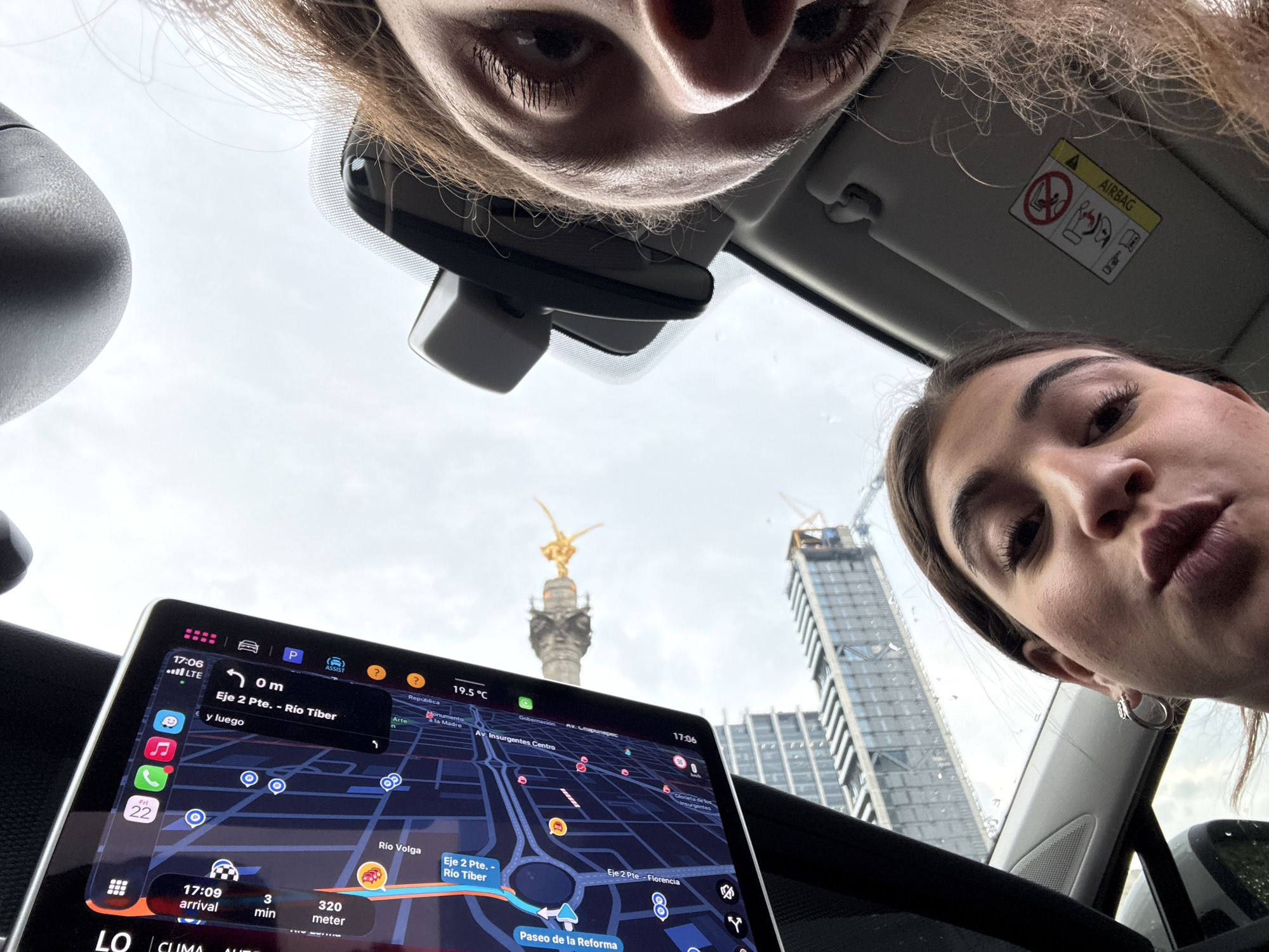 The image depicts two individuals in a car, presumably taking a selfie. The upper figure is cropped closely, showing the upper portion of their face, including eyes and nose. The other figure is positioned on the right, similarly cropped, focusing on their face from the nose down to the chin.
The car's interior is visible, with a touchscreen display showing a navigation map. The map indicates the location as Mexico City, Mexico, specifically near "Eje 2 Pte. - Río Tíber" and "Paseo de la Reforma." The GPS shows "3 min" and "320 meter" to arrival. The screen also displays the current time as 17:06. The temperature is 19.5°C.
Outside the car window, the iconic "Monumento a la Independencia" (Angel of Independence) is visible, along with a modern building under construction. The sky is overcast. Other elements visible are the car's rearview mirror, sun visor with an airbag warning label, and dashboard.