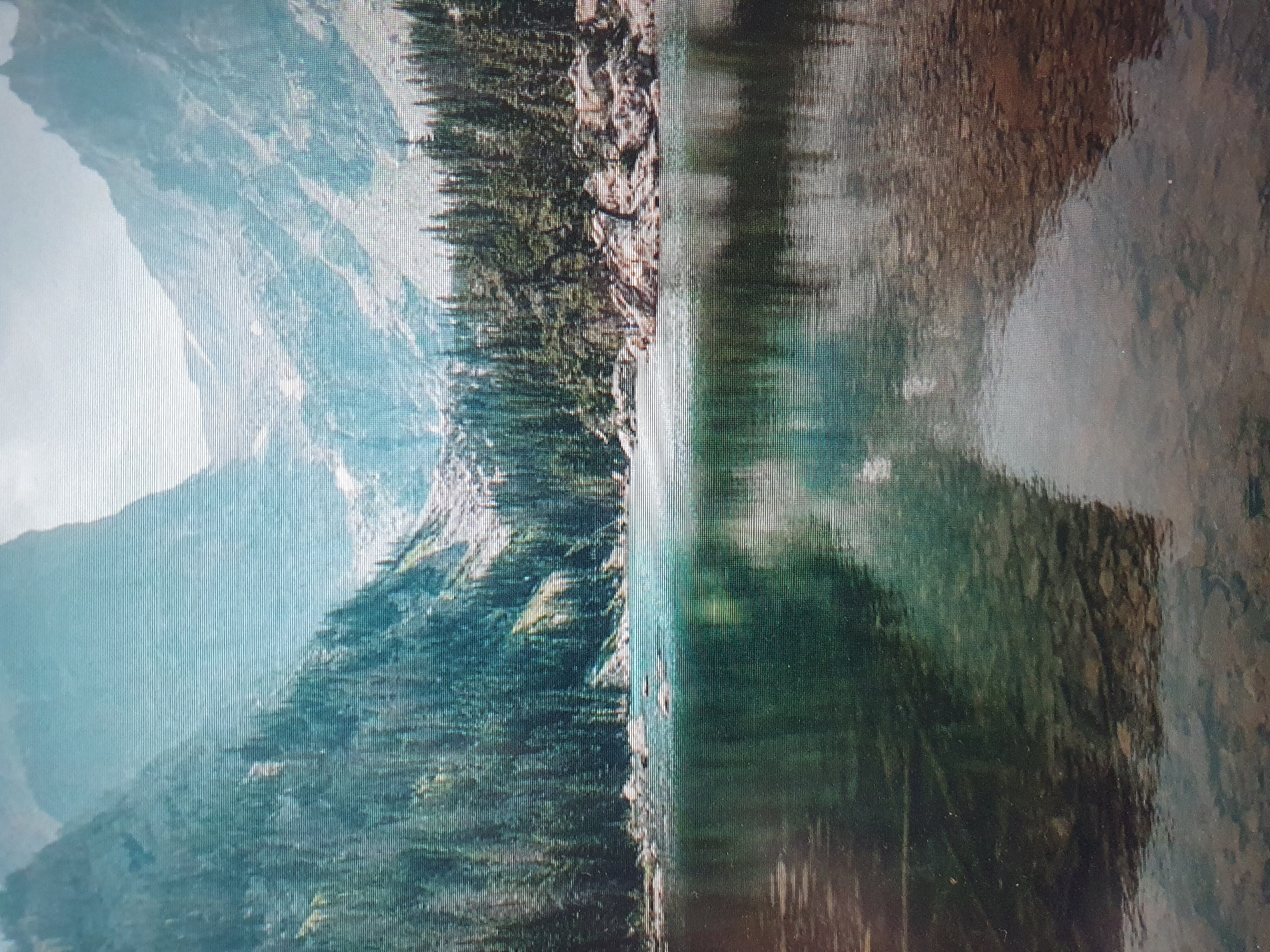 The image depicts a serene mountain lake scene, with towering, mist-shrouded mountains in the background. A dense evergreen forest lines the shores of the lake, and the calm water reflects the surrounding landscape. The foreground shows the clear, rocky bottom of the lake. The scene is natural and undisturbed. There are no people, man-made objects, or any indication of urban environments like Tashkent, Uzbekistan.