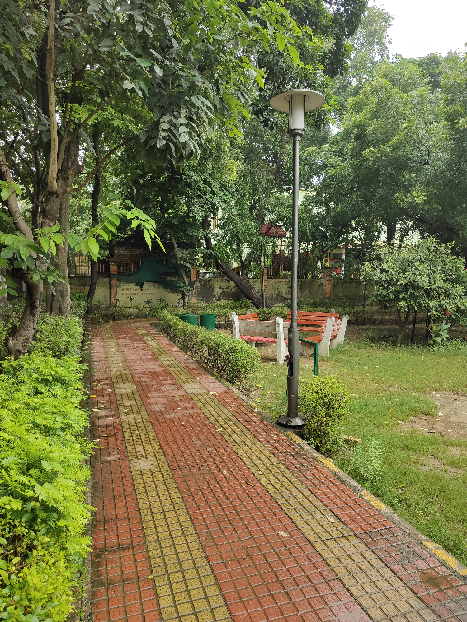 The image depicts a paved pathway in a park in Gurgaon, India. The pathway is made of red and yellow square tiles and is wet, suggesting it might have recently rained. Lush green bushes line the left side of the path, and manicured hedges border the right side. In the background, there are several park benches with red seating and white supports, a lamppost, and various trees. A wall with some writing and drawings is visible behind the benches. The overall environment appears to be a well-maintained urban park.