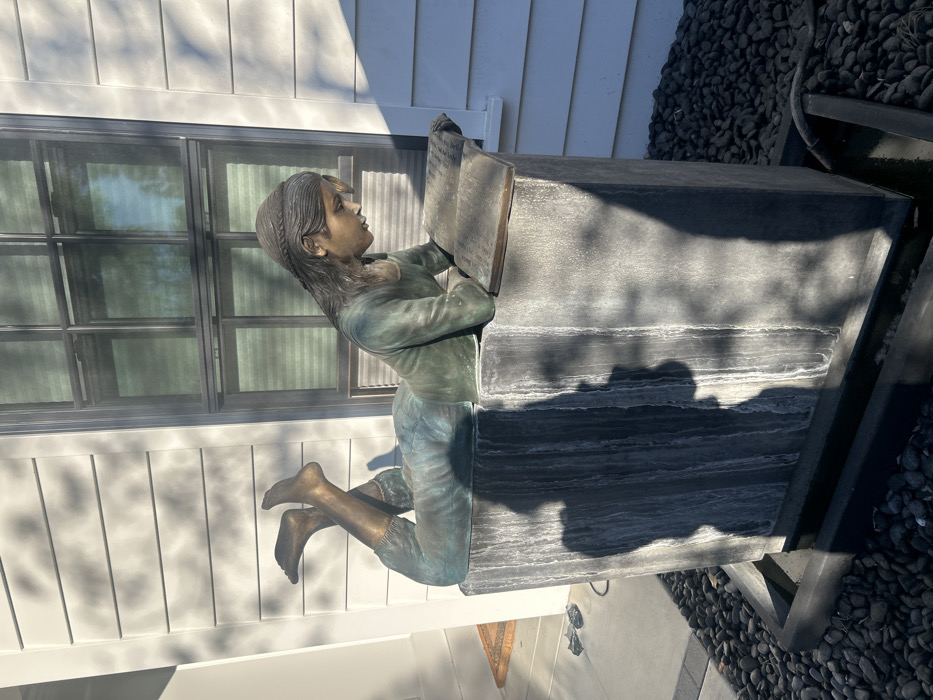 A bronze statue of a girl is reading a book atop a stone bench. The girl has long hair and wears casual clothes. The bench is positioned in front of a white house with a window. Dark pebbles surround the base of the bench. The statue appears to be in an outdoor setting. The sun casts shadows across the house and bench.