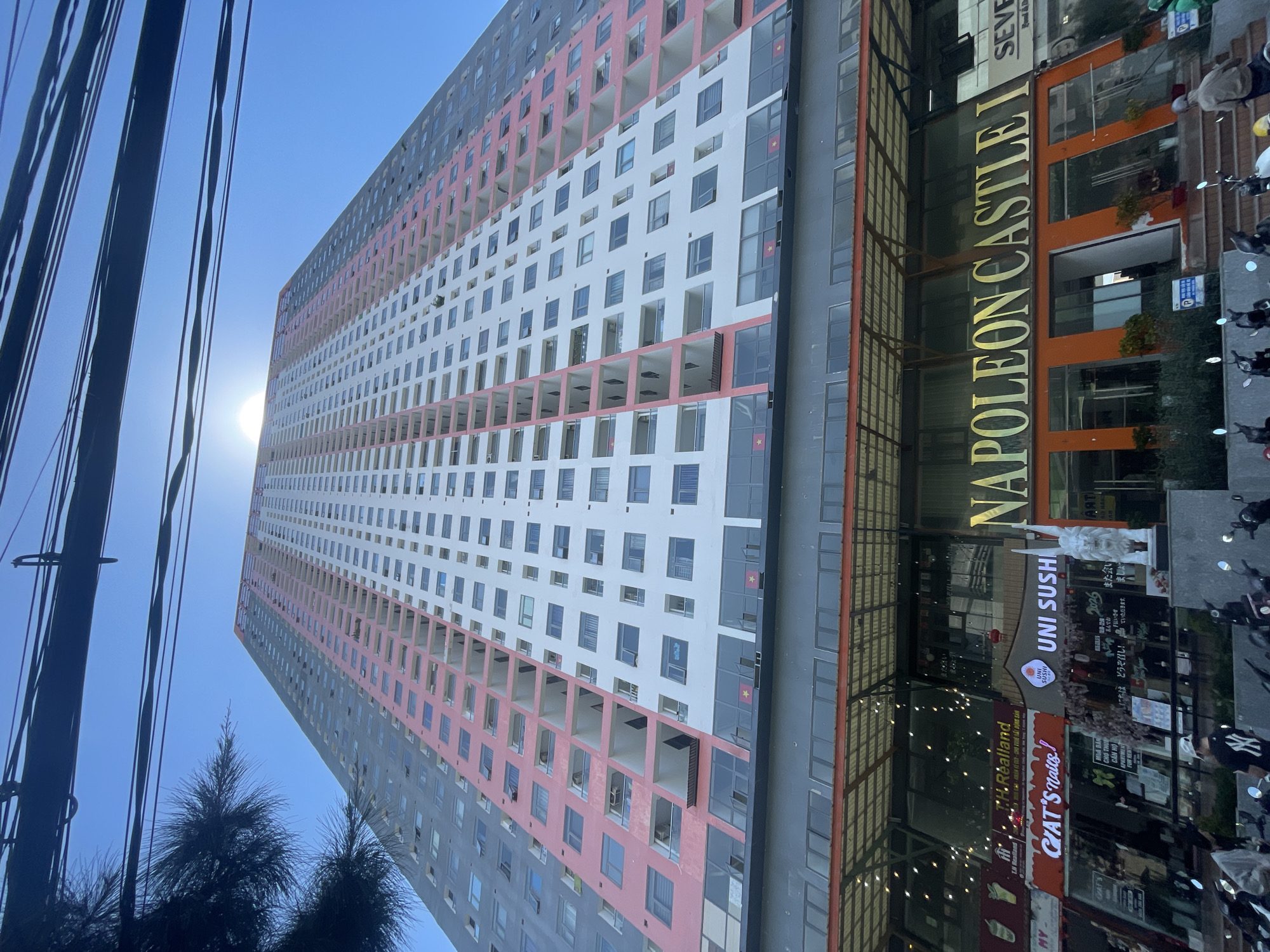 An extreme wide-angle, low-angle shot captures the Napoleon Castle I residential building in Bac Nha Trang Ward, Vietnam. The towering structure dominates the frame, its facade a mix of white, pink, and grey sections. Numerous balconies with white railings are visible, interspersed with rows of windows. The sun shines brightly, creating a lens flare in the upper portion of the sky. Several power lines are strung across the top of the image, and the dark branches of a tree are visible in the upper left corner.
At the base of the building, commercial establishments are visible, including a sign for "NAPOLEON CASTLE I" in large gold lettering above an orange facade. Below this sign, several storefronts are apparent, featuring signs for "CAT'S Nails," "UNI SUSHI," and "SEVEN Food & Drink." In front of the stores, there are rows of parked motorcycles and a few people, one wearing a baseball cap and a dark t-shirt with a white logo, and another wearing a light-colored jacket and a face covering. Another person in the background is walking away from the camera. The ground level appears to be a paved walkway with some potted plants. The image is taken during the day under a clear blue sky.