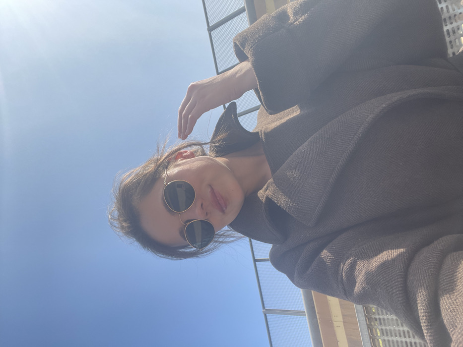 A female individual is depicted in a close-up, low-angle outdoor shot under a clear blue sky in Rome, Italy. The subject wears dark, round sunglasses with thin gold frames and a dark brown, possibly wool or tweed, overcoat with a large collar. Her dark hair is pulled back, with some strands appearing loose around her face. Her right hand is raised to the right side of her head, with fingers slightly splayed, appearing to be near her temple or hair. A subtle smile is present on her lips, which have a glossy appearance. Strong sunlight illuminates the scene, creating a lens flare in the upper right quadrant of the image. In the background, a metal railing or fence with horizontal bars is visible, suggesting an elevated outdoor area such as a bridge or terrace. A light-colored structure is partially visible behind the railing.