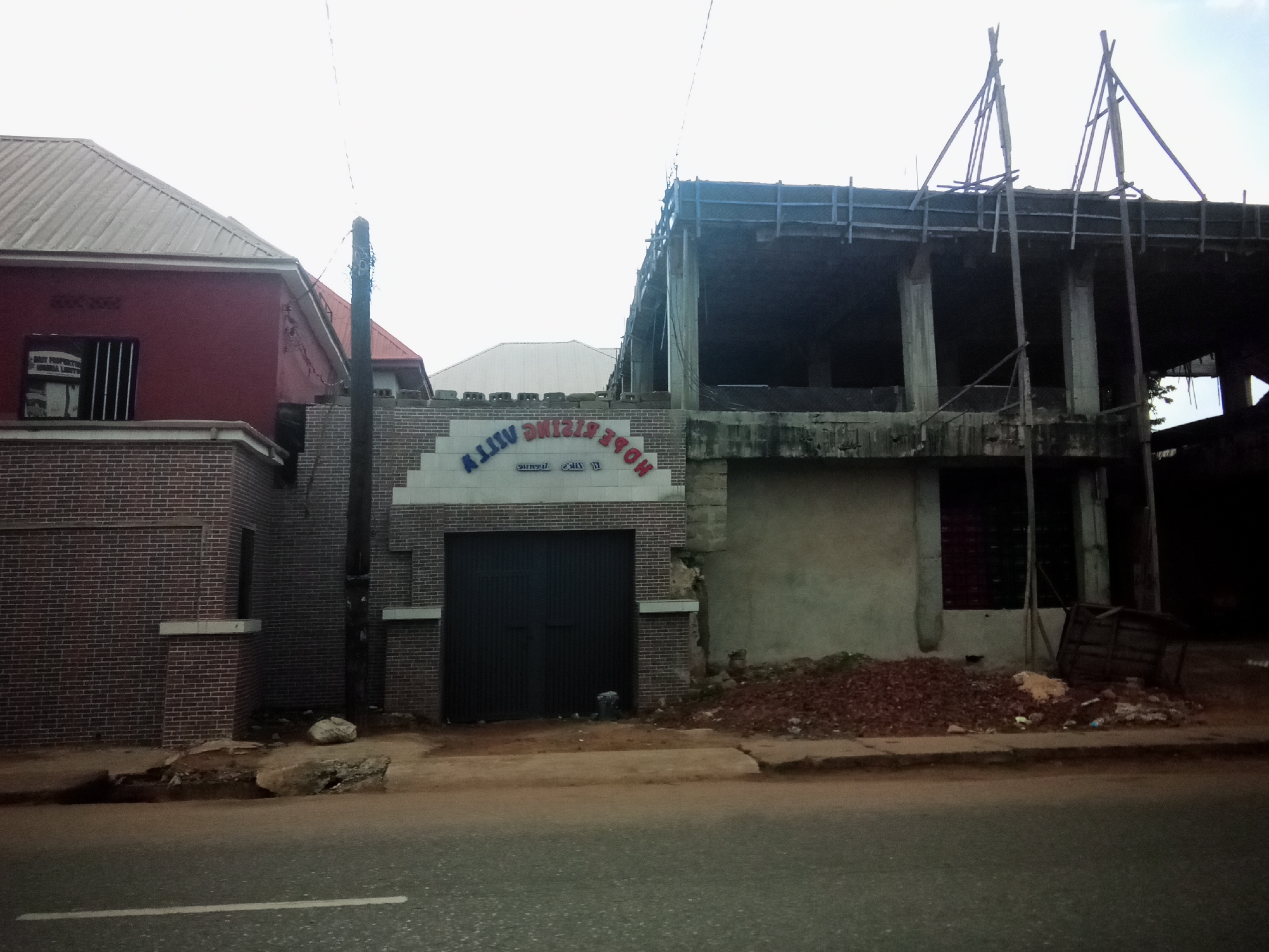 The image depicts a street scene in Nibo, Nigeria, featuring a brick building with a red facade on the left and a multi-story building under construction on the right. The brick building has a corrugated metal roof and a barred window. Adjacent to it is a smaller building with a grey brick facade and a large dark metal garage door. Above the garage door, a sign reads "HOPE RISING VILLA" in multi-colored lettering, with some letters appearing reversed. The building under construction is a concrete structure with exposed rebar and wooden scaffolding on the upper levels. Piles of reddish-brown dirt and debris are visible on the ground in front of both buildings, and the street in the foreground has a white painted line. A dark-colored utility pole stands between the two buildings. The sky is overcast.