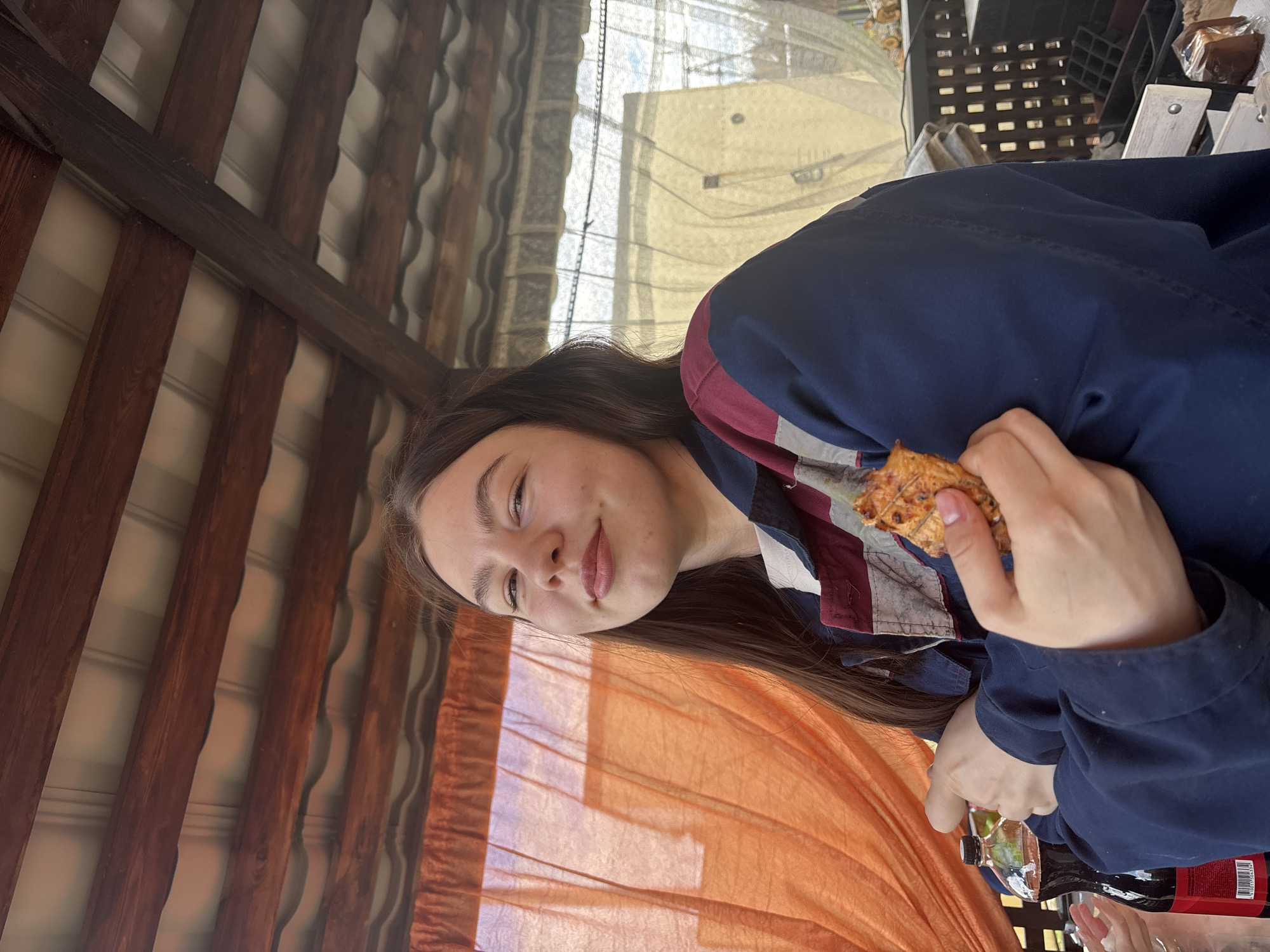 A young woman is seen in a medium shot, holding a piece of food, possibly a small pizza, in her right hand. She is wearing a dark blue jacket over a collared shirt with stripes and patterns, while facing the camera with a slight smile. Her long brown hair falls down her shoulders. Behind her, there is an orange curtain or fabric. The background features a wooden-beam structure, possibly a roof or pergola, and through the opening, a curtained window and a metal chair can be seen. There are other objects visible on the table in front of her, including bottles and wrappers. The location of the city of Chaikovsky, Russia cannot be determined from this image.