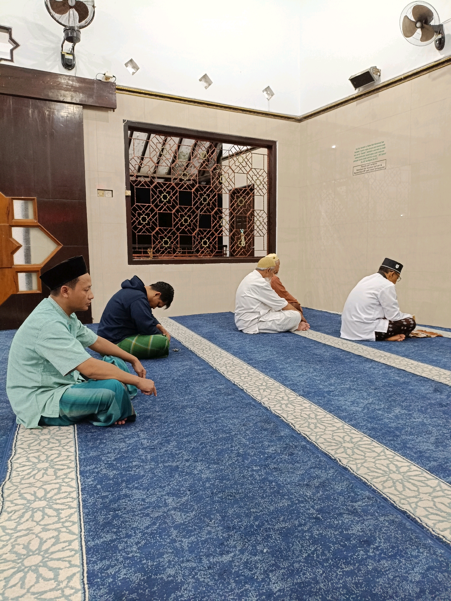 Four men are seated on a blue mosque carpet. The man in the foreground is wearing a light green shirt, a black cap, and a sarong. He is sitting cross-legged with his right hand resting on his knee. To his left, a younger man in a dark blue hoodie and a green sarong is kneeling with his head bowed. Further back, two older men are also seated on the carpet. One is wearing a white robe and a cream-colored cap, while the other is in a white robe and a black cap, facing away from the camera and appearing to read a book. The room has white tiled walls, a dark wooden door on the left, and a patterned window with dark wooden framing on the back wall. A ceiling fan is visible in the upper left corner, and two lights are mounted on the ceiling. The carpet has a decorative white and blue pattern running in parallel lines. The scene appears to be in a mosque in Maguwoharjo, Indonesia, likely during prayer.