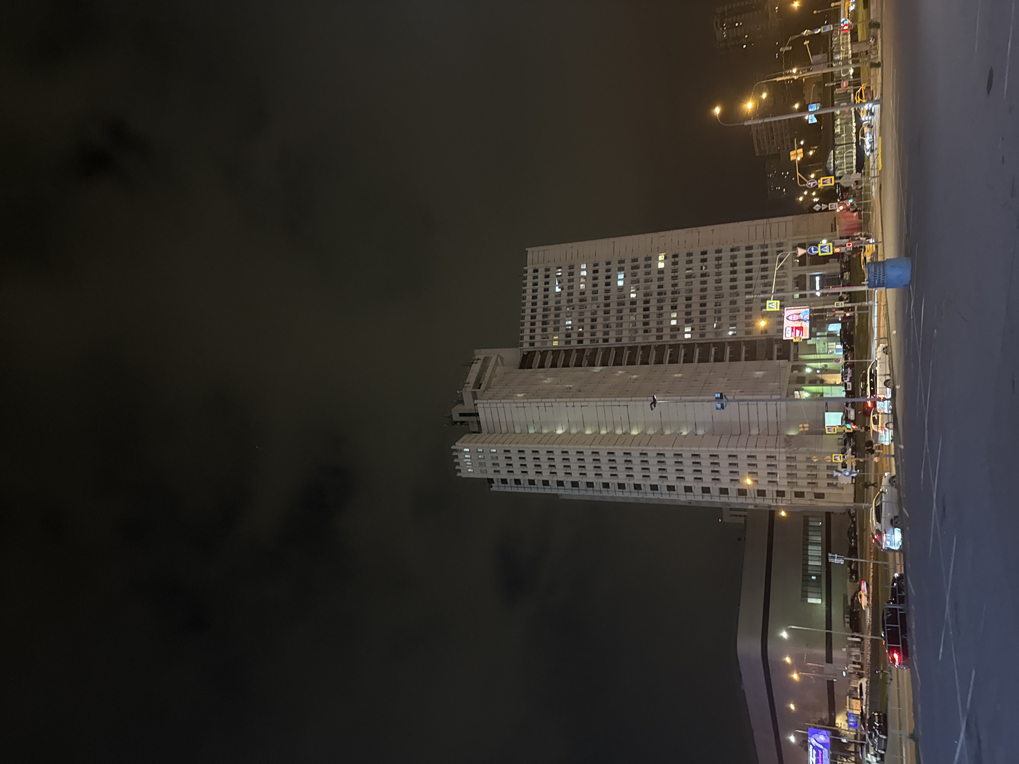 The image shows a tall, white, rectangular building at night in Moscow, Russia. The building has many windows and appears to be either an office or residential building. A paved area is adjacent to the building, and several street lights illuminate the area, as well as the building. There are other buildings in the background, and the sky is dark. Some traffic signs are near the roadway.