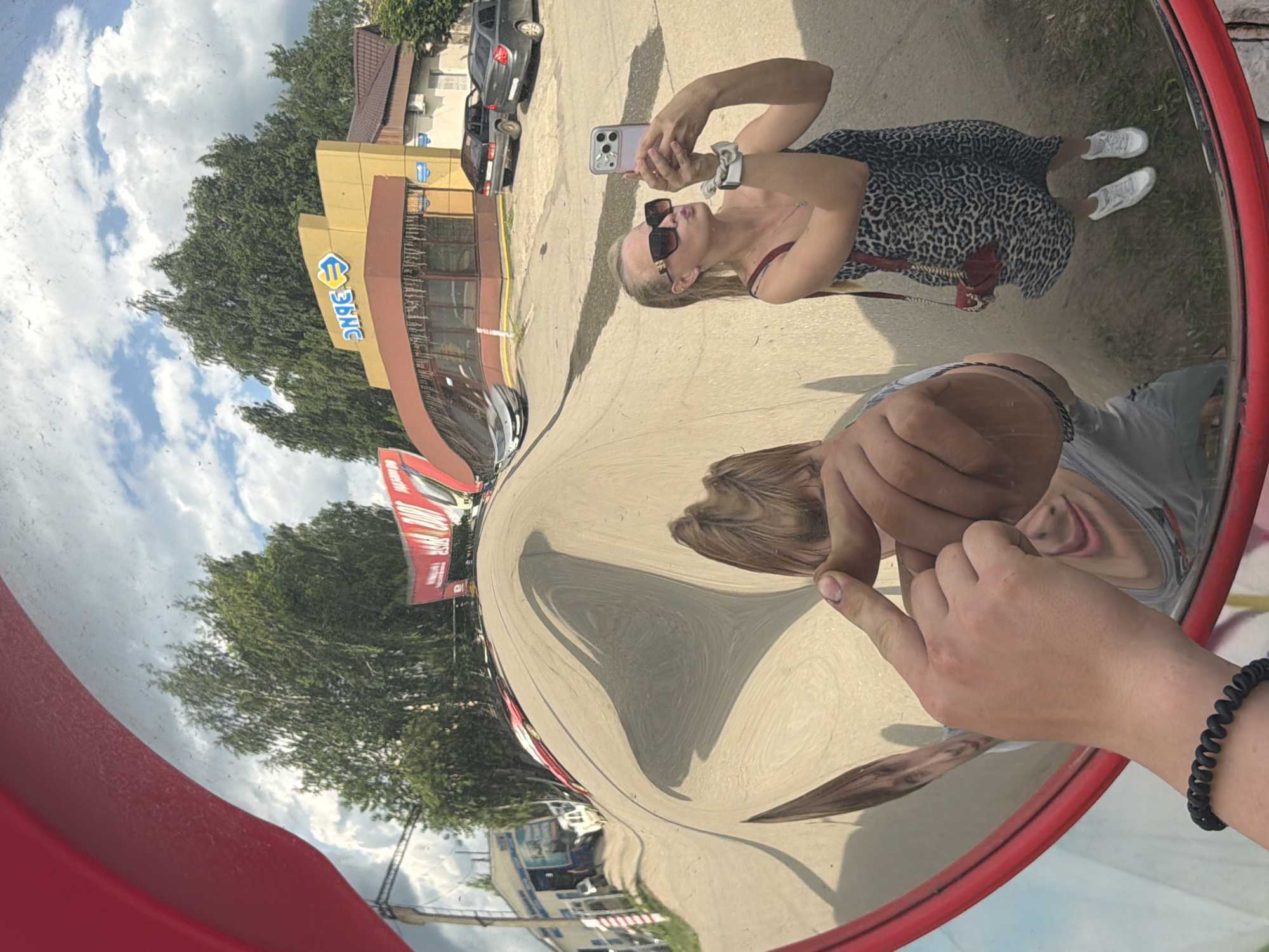 The image shows a wide-angle reflection in a convex mirror. In the reflection, a woman is taking a selfie with her phone. She is wearing a leopard-print dress, sunglasses, and white sneakers. She has a small red purse with a gold chain slung over her shoulder. To the left of the woman, the reflection of a person's hand is visible, with a finger pointing towards the mirror. In the background, there is a yellow building with a sign that reads "ЭЛЛС". There are trees and a cloudy sky reflected in the mirror as well. The mirror is framed by a red border. This location is identified as being in the city of Chaikovsky, Russia.