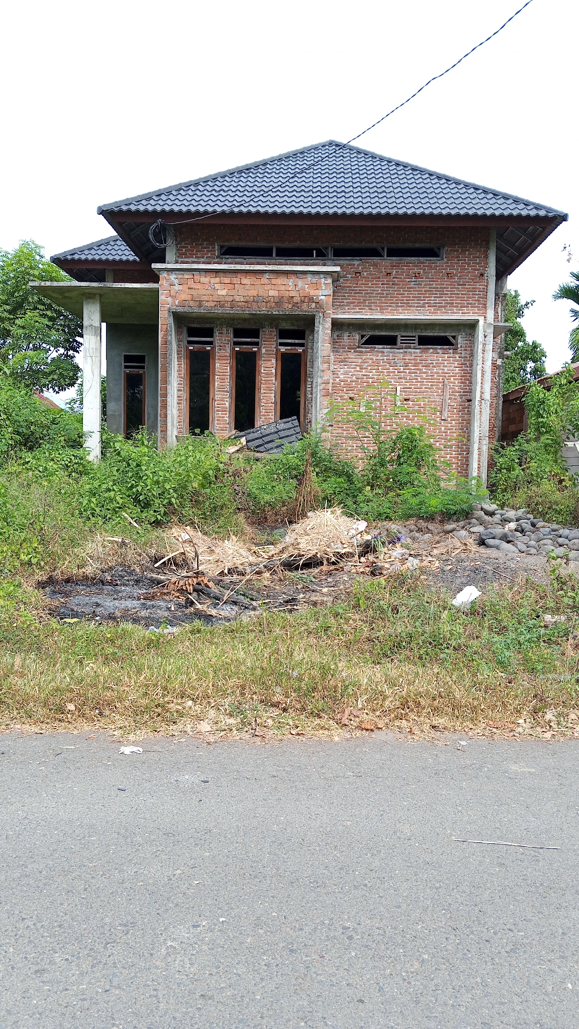 A partially constructed brick house stands in a semi-rural area, possibly Uteunreutoh, Indonesia. The house has a dark gray tiled roof and exposed brickwork. There are several large windows, some with dark wooden frames, and an unfinished concrete porch on the left. In front of the house, there is a pile of construction debris including straw, burnt wood, and rocks. Overgrown green vegetation surrounds the property. The foreground shows a paved road. The sky is overcast.