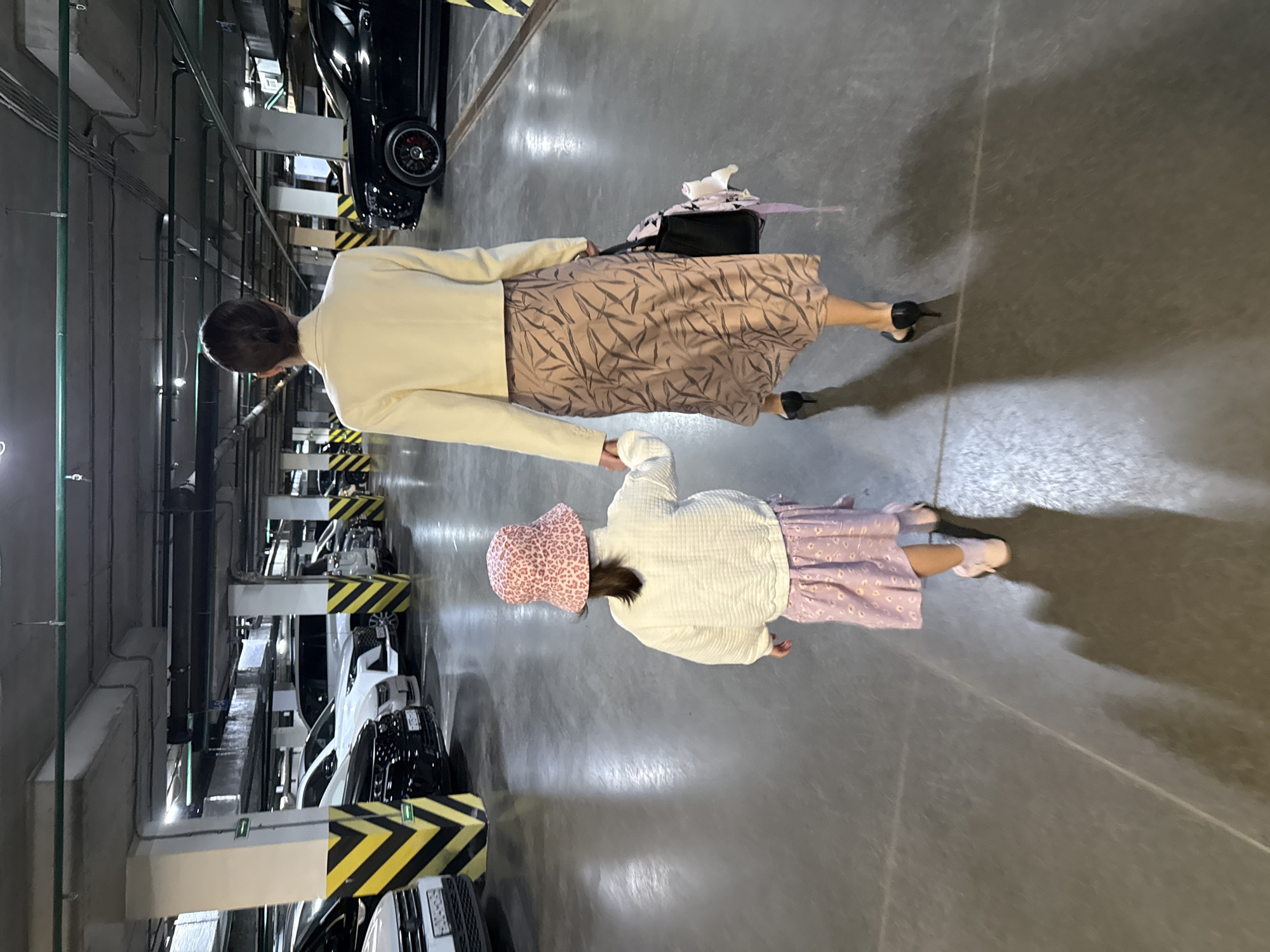 This image captures an adult and a child walking hand-in-hand through an underground parking garage. The adult, viewed from behind on the left, wears a light cream blazer, a light brown midi-skirt with a dark, abstract leaf-like pattern, and black high-heeled shoes. They carry a black shoulder bag. The child, also seen from behind on the right, wears a light-colored, textured long-sleeved top, a light purple skirt with a small white floral pattern, and light pink slip-on shoes. A pink bucket hat with a subtle leopard print covers the child's head.
The setting is clearly an artificially lit parking facility, with concrete floors, walls, and ceilings. Numerous cars, primarily black and white, are parked in stalls on both sides of the walkway. Yellow and black striped safety barriers mark the parking areas and structural columns. Exposed pipes and electrical conduits are visible along the walls and ceiling, typical of industrial or utility areas within such a structure. No specific time of day is indicated, as the environment relies entirely on artificial lighting. No discernible text or emotions are visible. The provided context indicates the location is Krasnoyarsk, Russia.