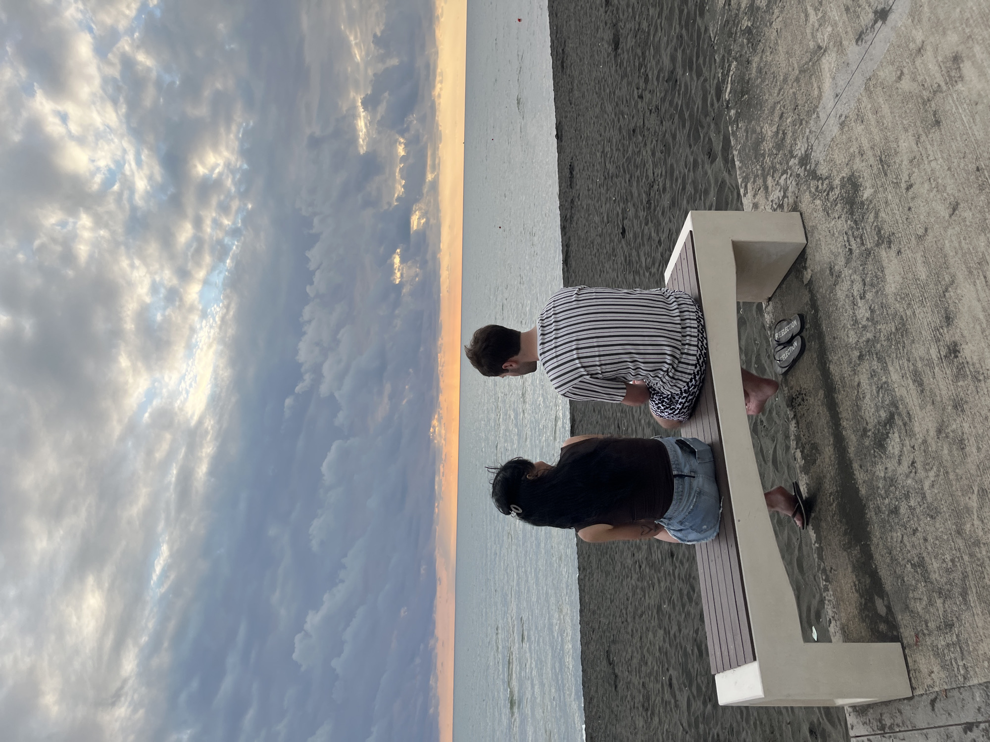 Two individuals, a woman and a man, are seated on a bench facing the sea in Ureki, Georgia. The woman, on the left, has long dark hair and is wearing a dark sleeveless top and a denim skirt. The man, on the right, has short brown hair and is wearing a vertically striped t-shirt and patterned shorts. They are sitting on a bench with a wooden slatted seat and a white base, positioned on a concrete path adjacent to a black sand beach. The sea stretches out before them, meeting the horizon where a gradient of orange and yellow hues suggests a sunset or sunrise. The sky above is filled with dramatic, textured clouds in shades of grey, white, and blue. The overall scene captures a moment of quiet contemplation against a backdrop of natural beauty.