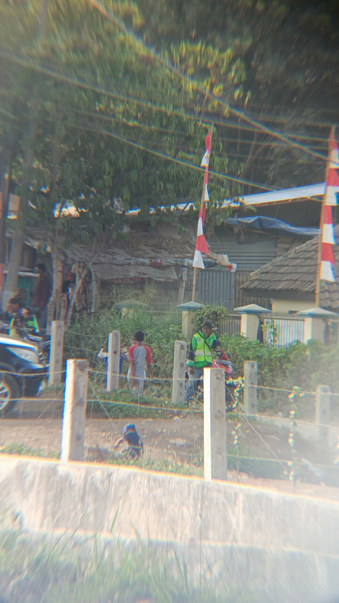 The image depicts an outdoor scene with several people and structures, likely in a semi-urban or rural setting. In the foreground, a gravelly path is visible with sparse grass. A low fence made of barbed wire and wooden posts separates this area from a grassy area beyond.
In the middle ground, several individuals are present. A person in a bright green vest and a dark cap is standing near a red motorcycle. To their left, a young boy wearing a gray and red t-shirt stands facing away from the camera. Further to the left, a dark-colored car is partially visible.
In the background, there are several structures. One appears to be a corrugated metal building, and another has a tiled roof. Indonesian flags, red and white, are displayed on poles, suggesting a national holiday or celebration. Trees and foliage are abundant throughout the scene, with some overhanging power lines. The lighting suggests it is daytime. The overall impression is of a community gathering or everyday life in a location that could be Margajaya, Indonesia, given the presence of Indonesian flags.