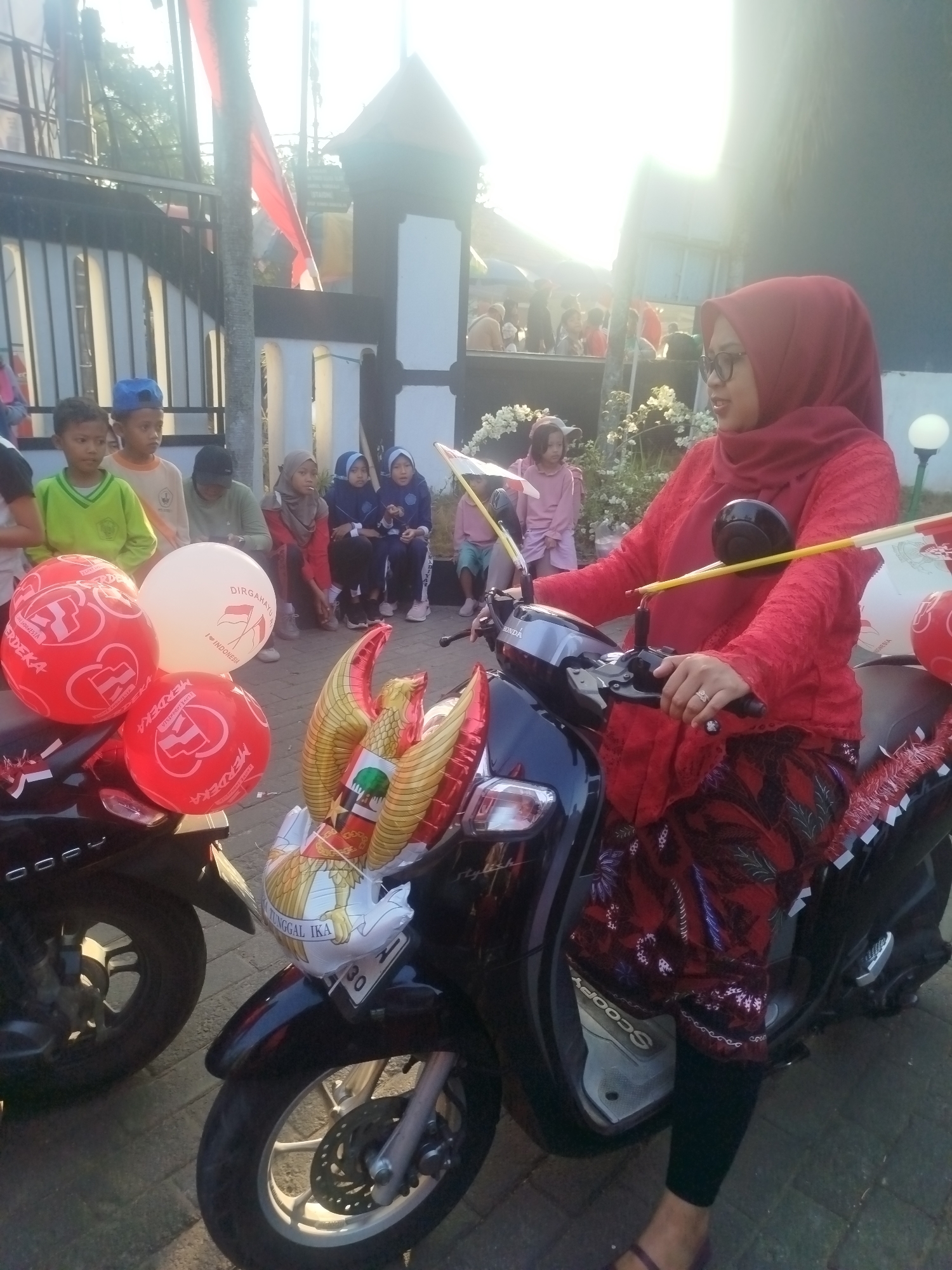 In Burneh, Indonesia, a woman wearing a red hijab and a red, lace-patterned top with a patterned red skirt sits on a black scooter. She holds the handlebars and is looking to her right. The scooter is decorated with several balloons, including two red balloons with white text and logos, and a large white balloon with red text and logos. In front of the woman, a Garuda Pancasila balloon is attached to the scooter. Behind her, a group of children are gathered, some wearing school uniforms. The background includes a building with a dark roof and white pillars, and a tree with bright sunlight filtering through its leaves. The scene appears to be outdoors, with a paved ground.